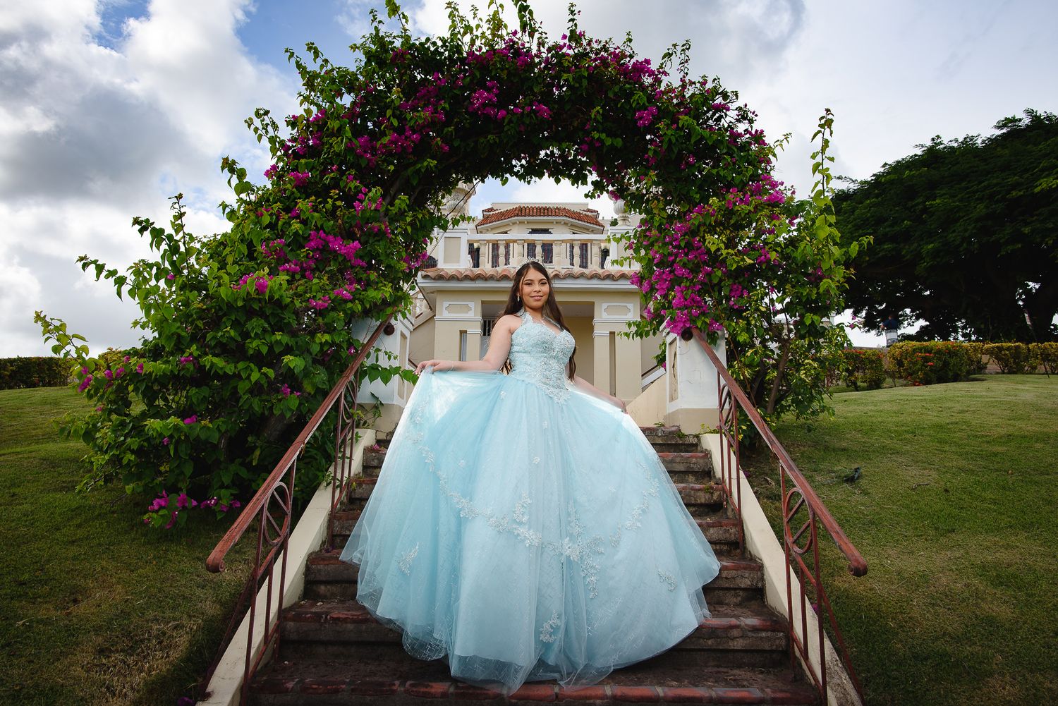 Young woman in a light blue ball gown stands under a floral arch with pink flowers at a elegant estate.