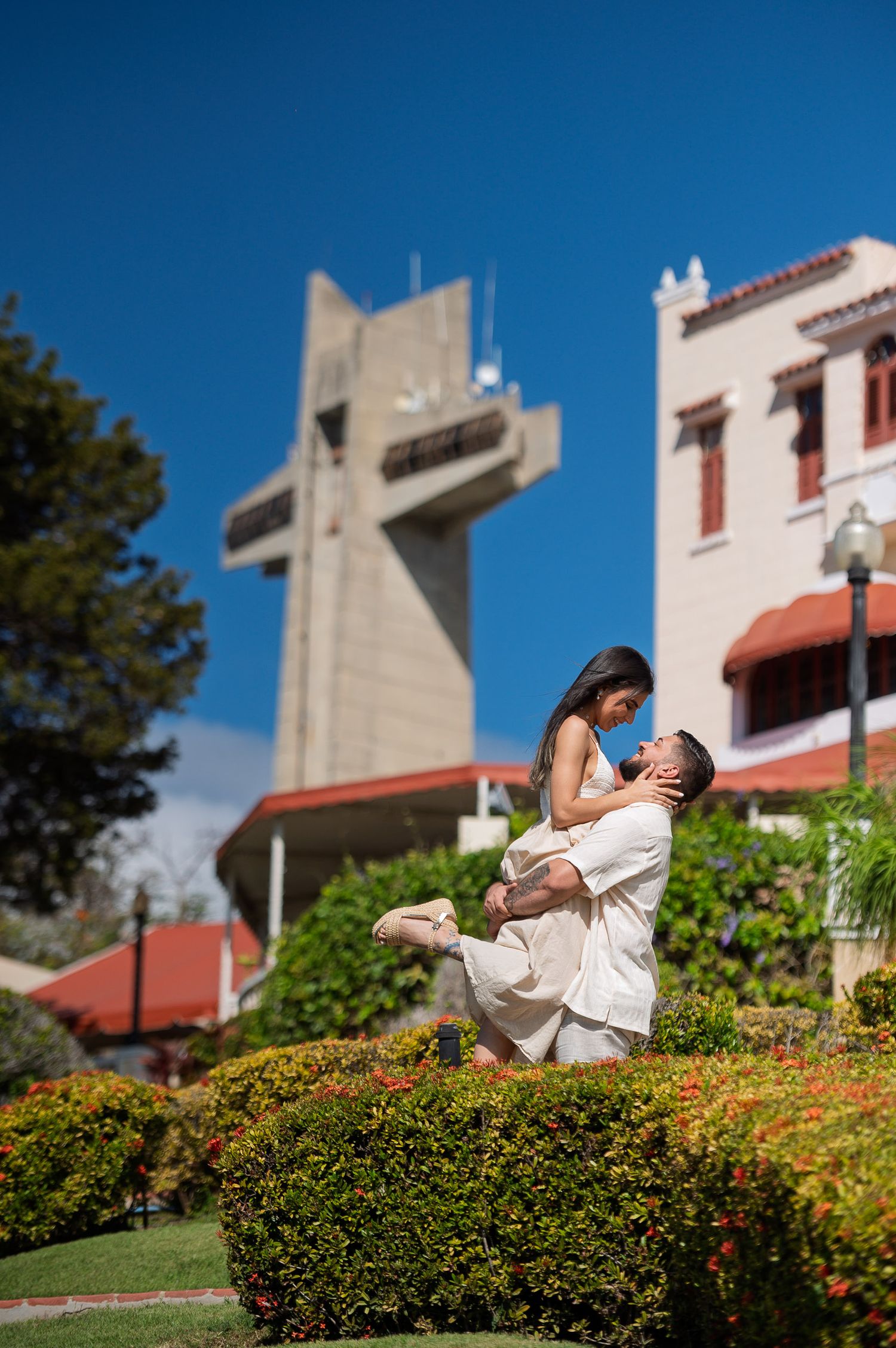 Couple embracing in garden with iconic cross monument and historic building under blue sky in Ponce, Puerto Rico.