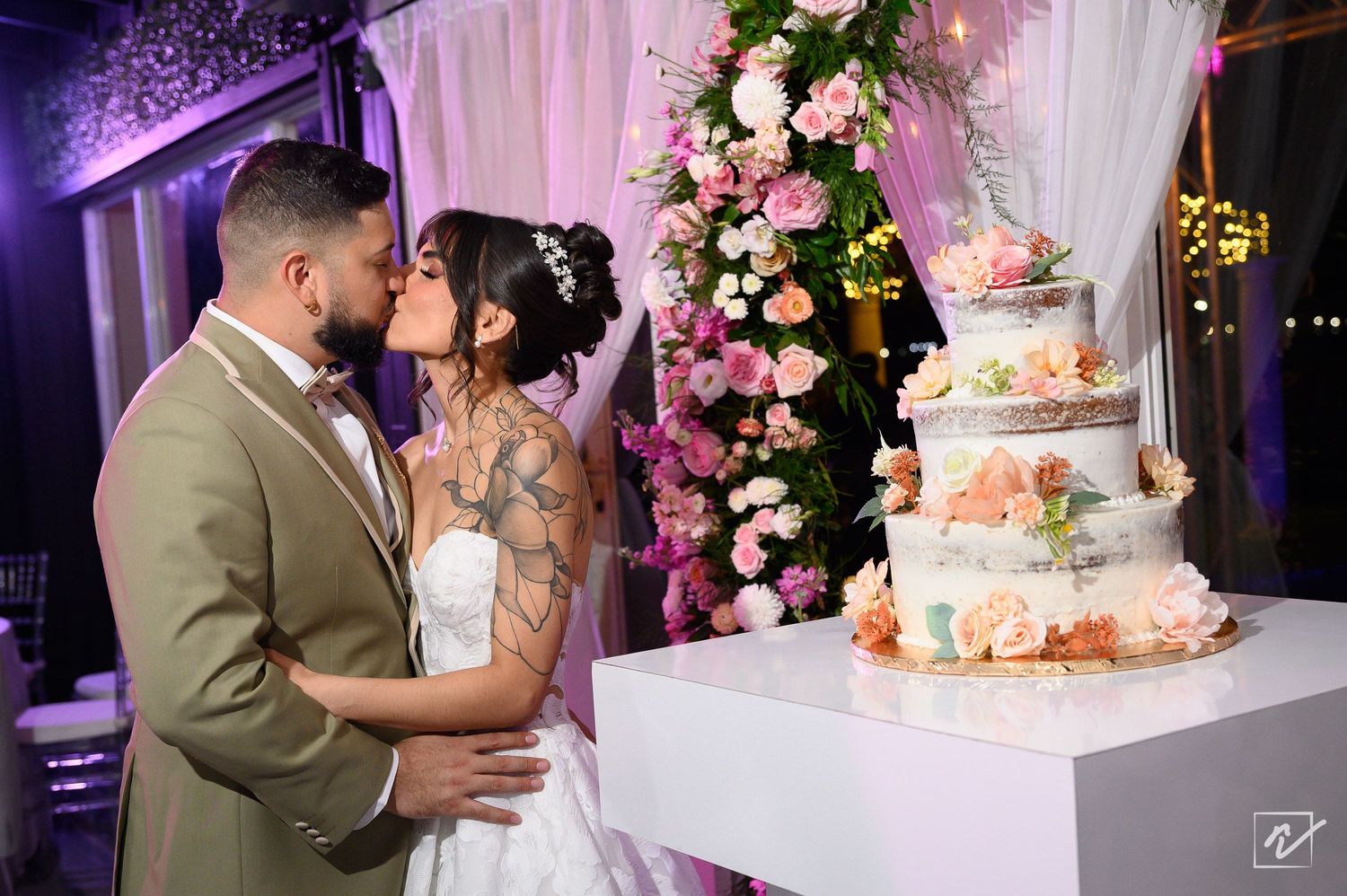 Bride and groom share a kiss beside a tiered wedding cake and lush pink and white floral arrangement at an elegant reception.