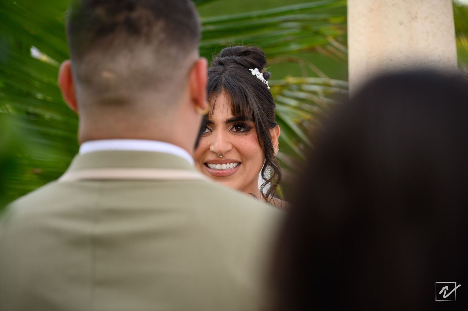 Smiling bride with a floral hair accessory looks past a groom in a tan suit during an outdoor tropical wedding ceremony.