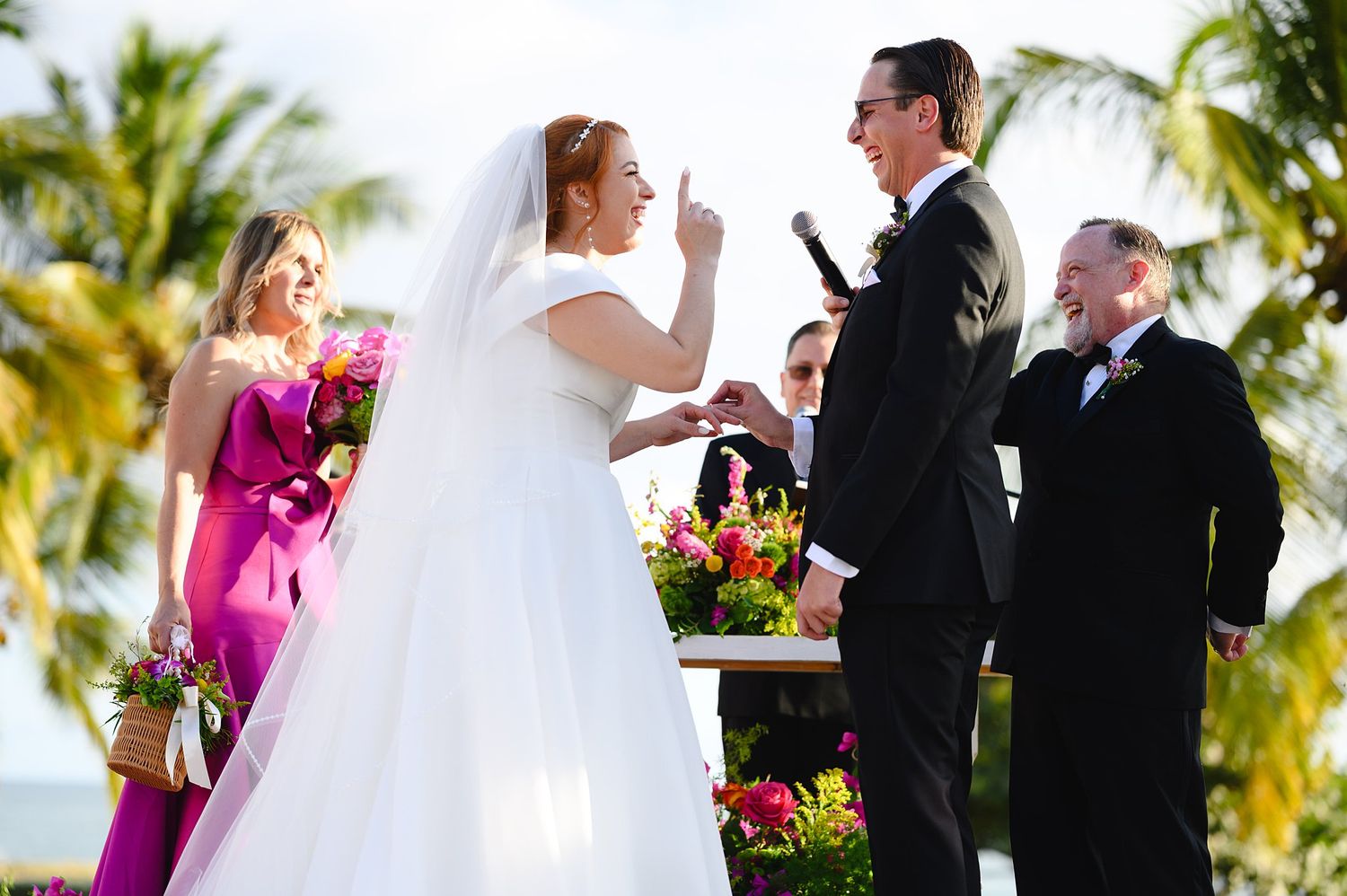 Bride and groom exchange vows at outdoor beach wedding ceremony with tropical palm trees and pink floral arrangements.