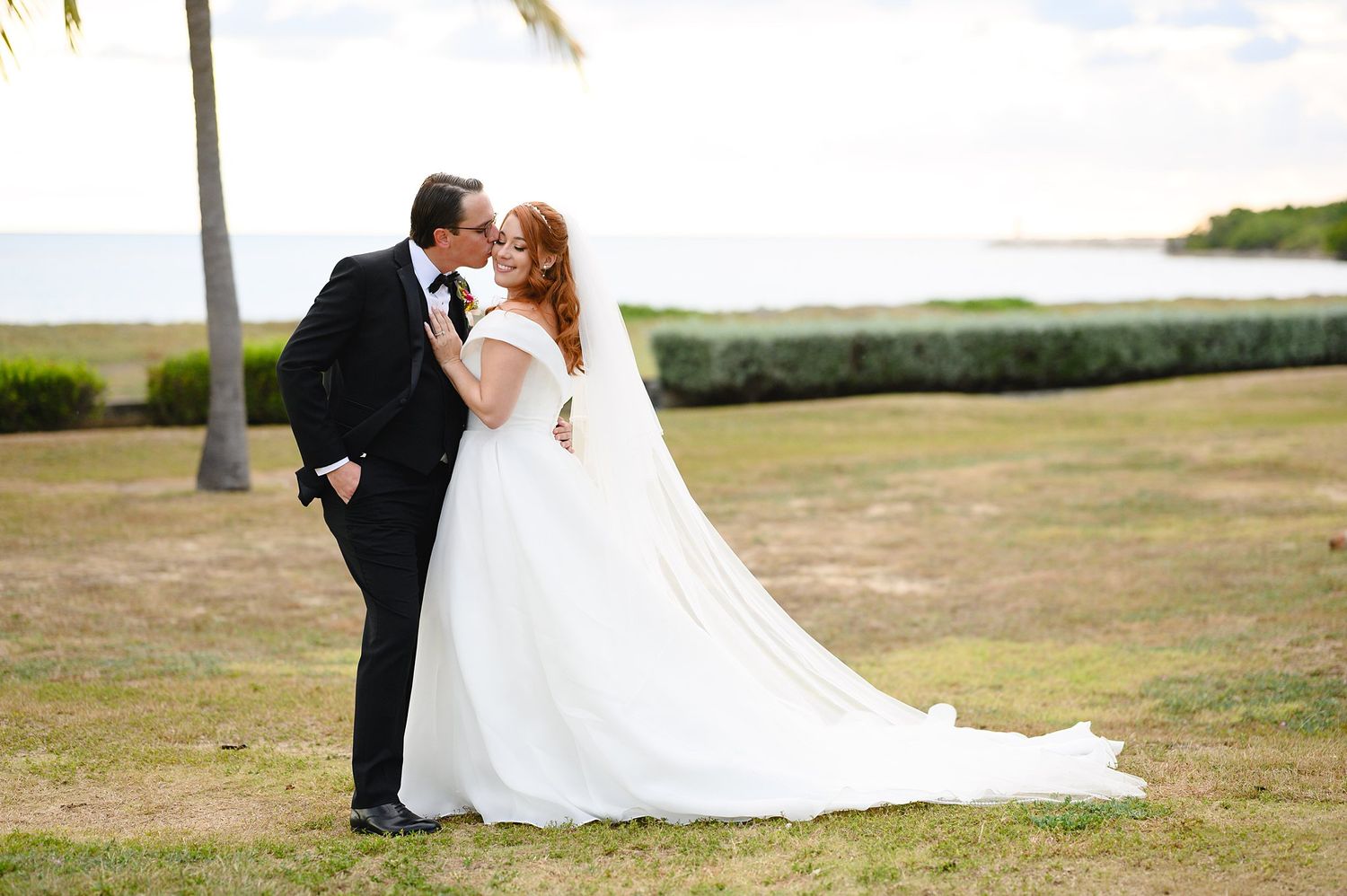 Bride and groom share a romantic kiss on a tropical lawn near the ocean, with a palm tree and lush greenery in the background.