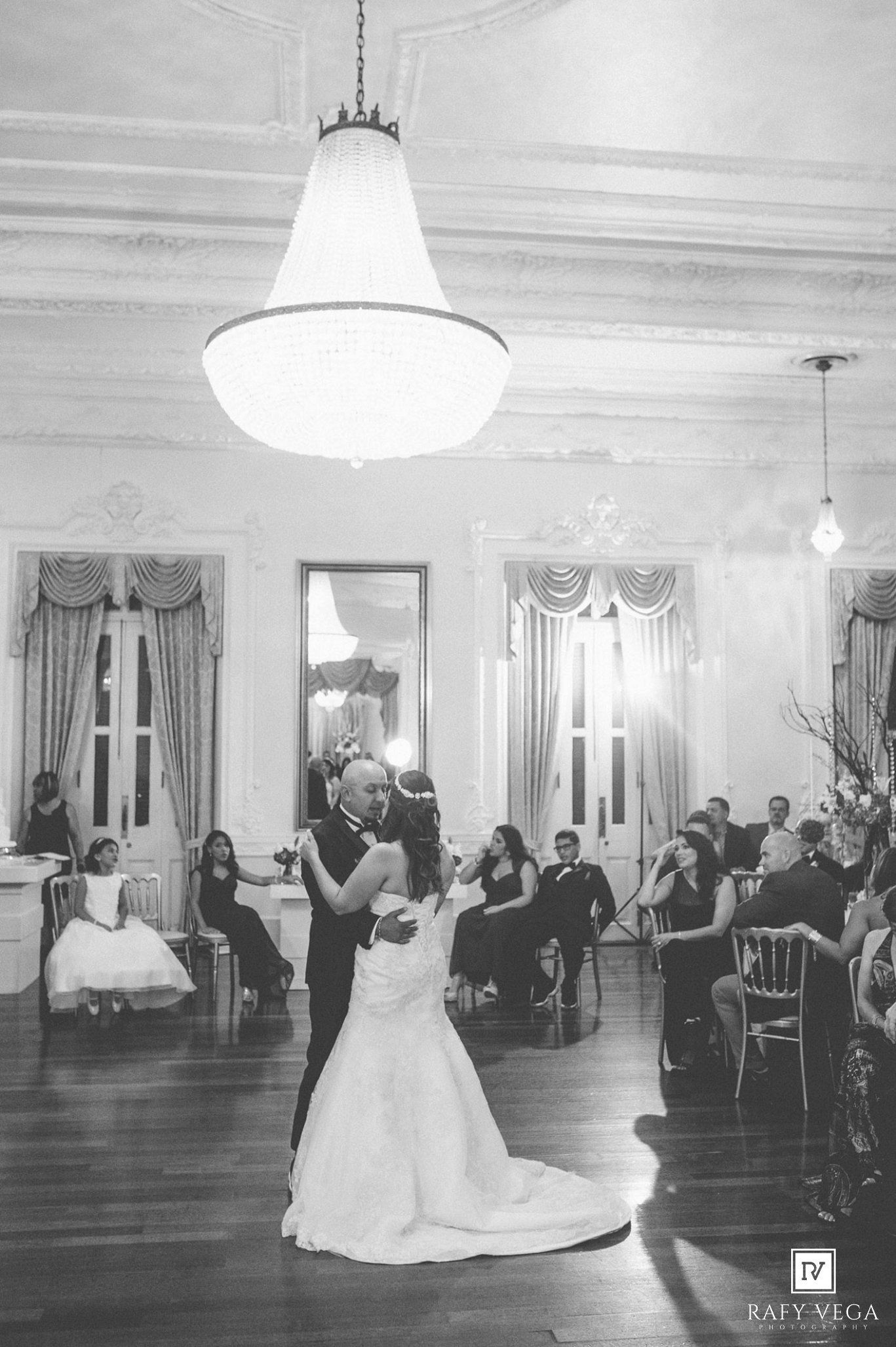 Black and white photo of bride and groom sharing first dance at elegant wedding reception with chandelier.