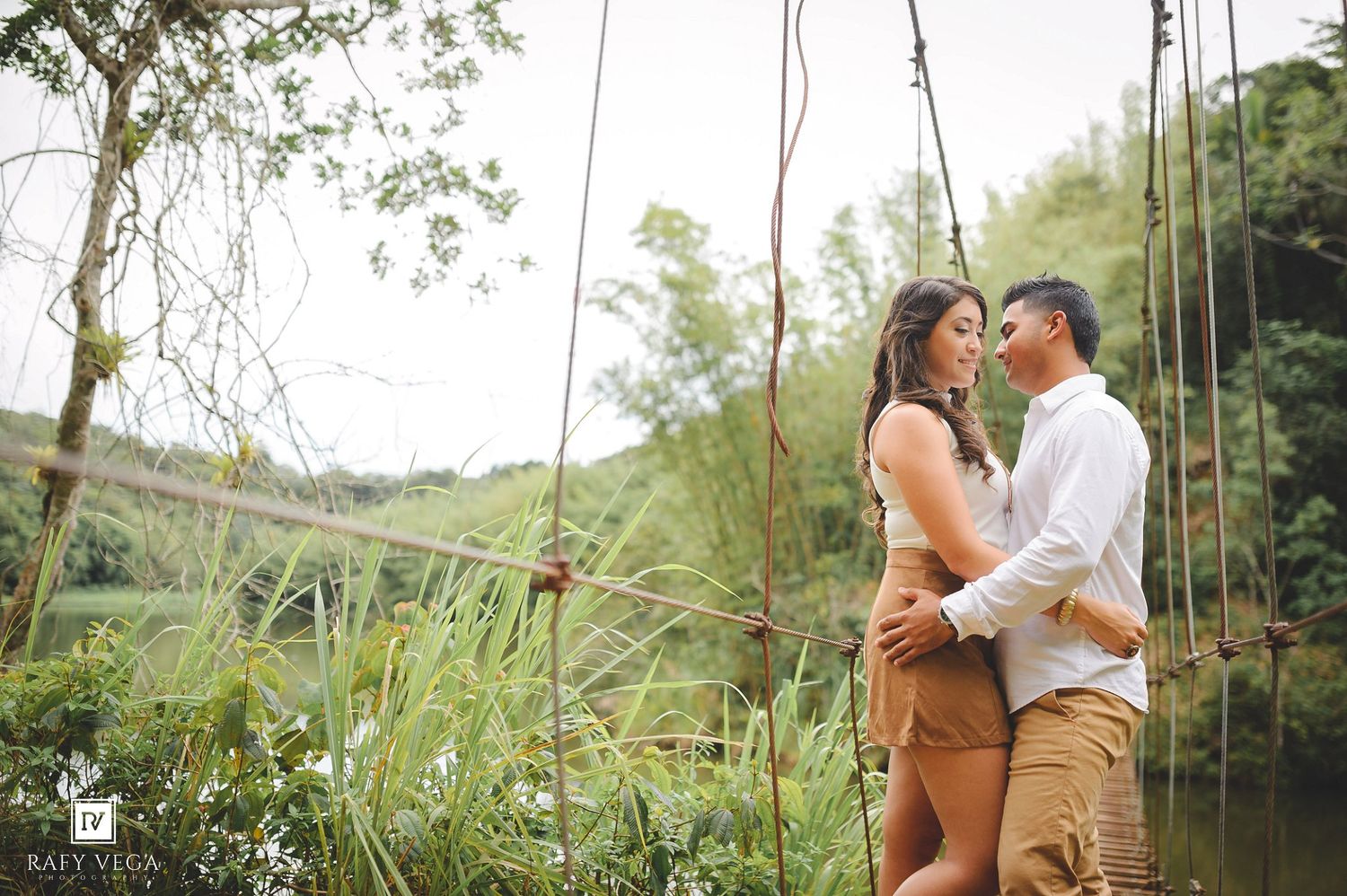 Couple embracing on a rustic rope bridge surrounded by lush greenery and a serene lake in a romantic outdoor setting.