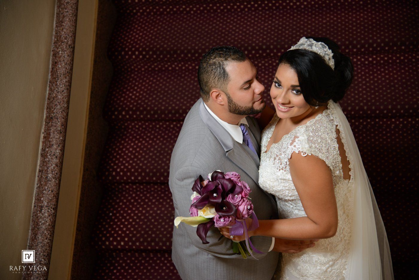 Bride and groom share a romantic moment on staircase, bride holding purple calla lily bouquet in white lace gown.