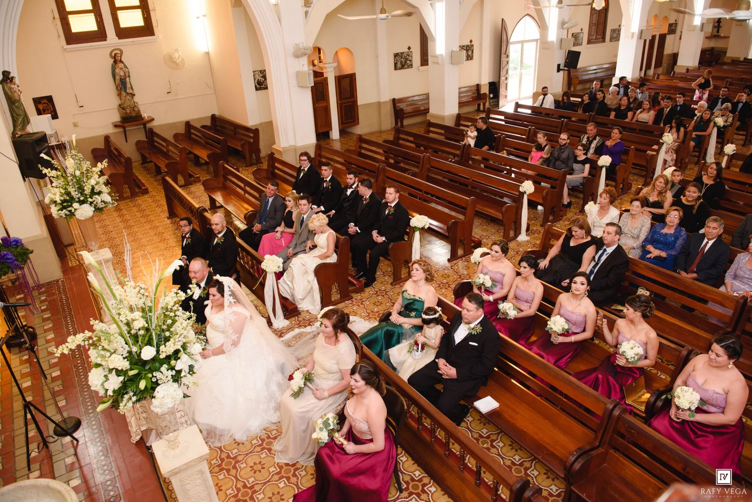 Aerial view of a wedding ceremony in a church with guests seated in wooden pews and bridesmaids in pink dresses.