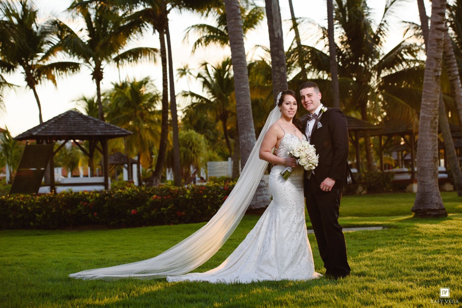 Bride and groom pose on lush tropical lawn with palm trees and gazebo at golden hour wedding venue.