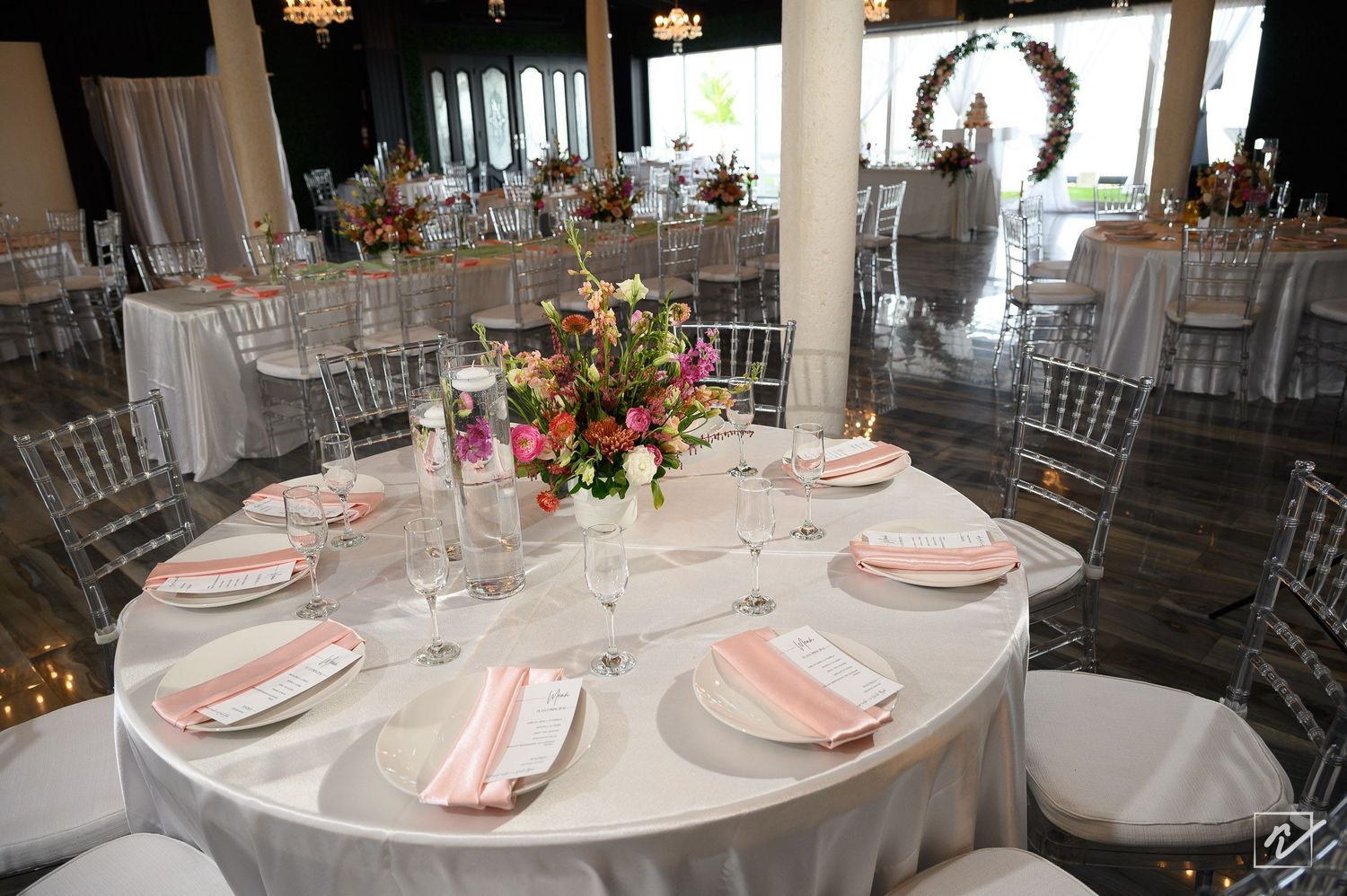 Elegant wedding reception table with pink napkins, white plates, and a fresh floral centerpiece in a bright banquet hall with round tables.