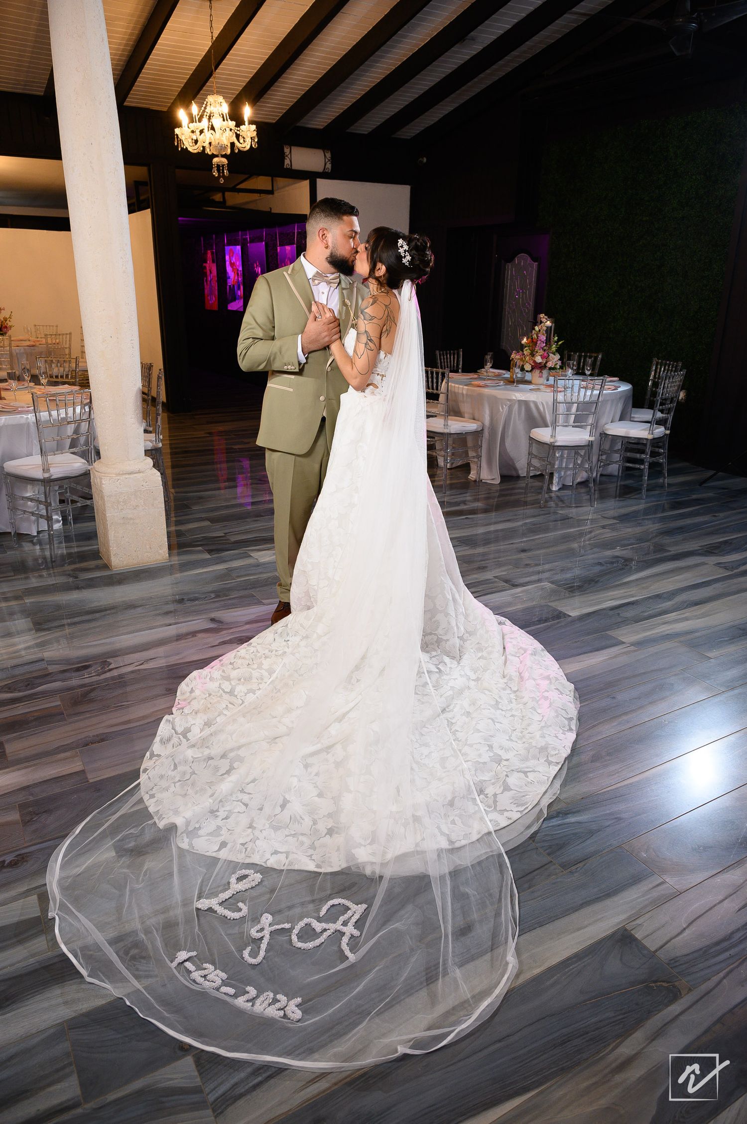 Bride in a lace gown with a long monogrammed veil embraces her groom in an olive suit in an elegant wedding reception hall.