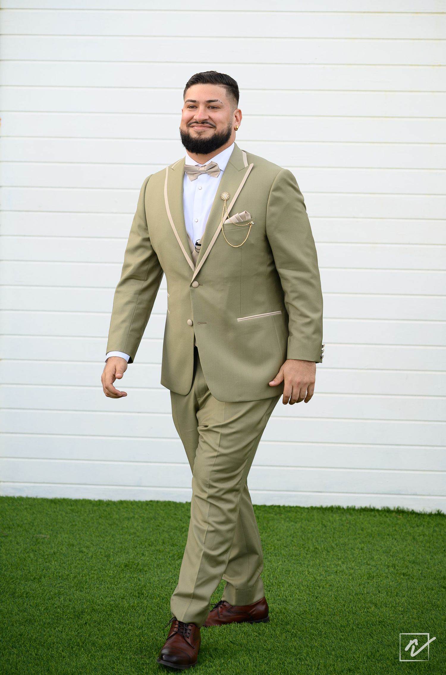 Smiling groom with a beard walks across artificial grass in a sage green suit with a bow tie and pocket square before a white wall.