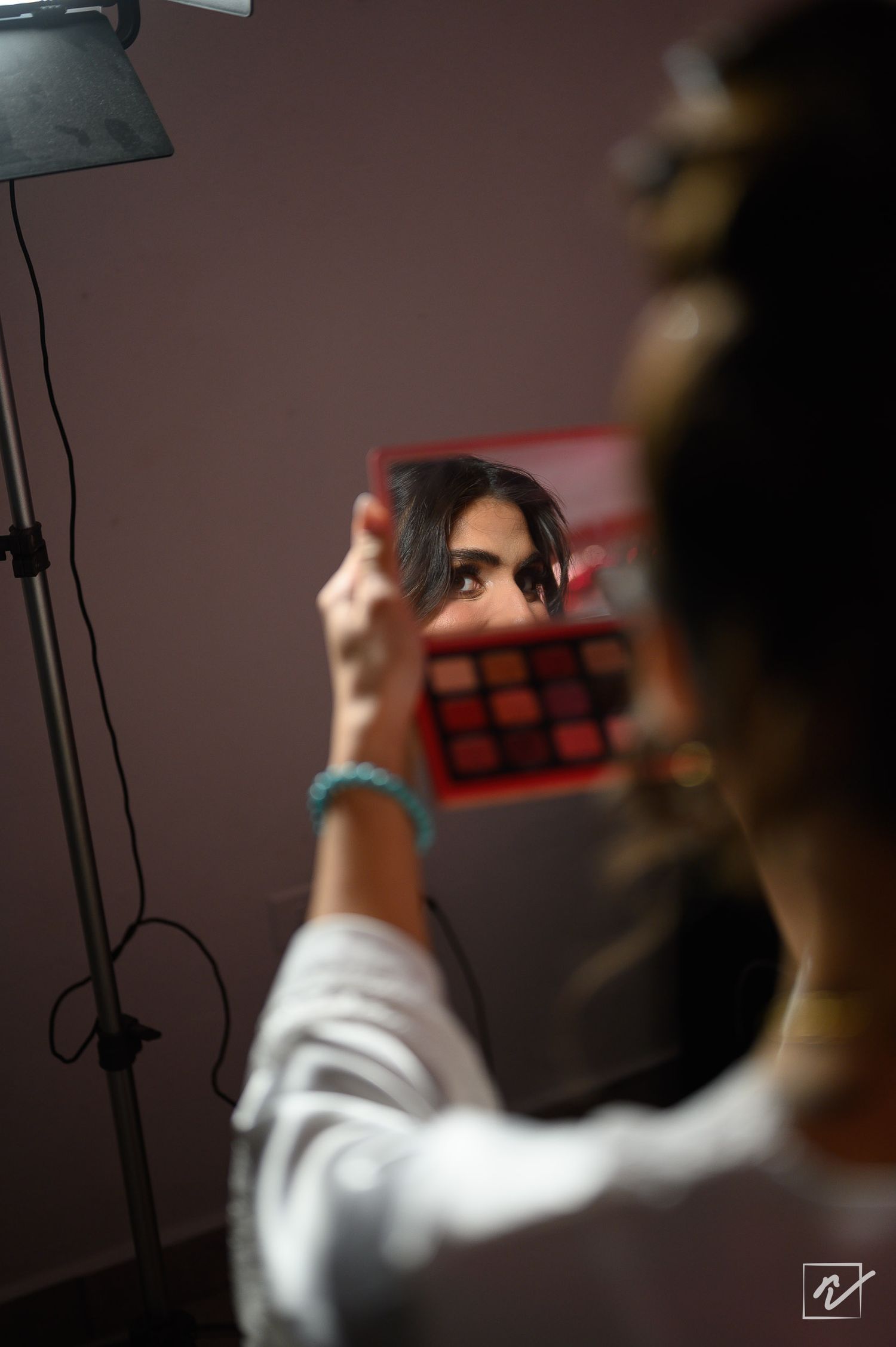 Woman in a white blouse holds a small mirror showing her reflection beside an eyeshadow palette while getting ready near a studio light.