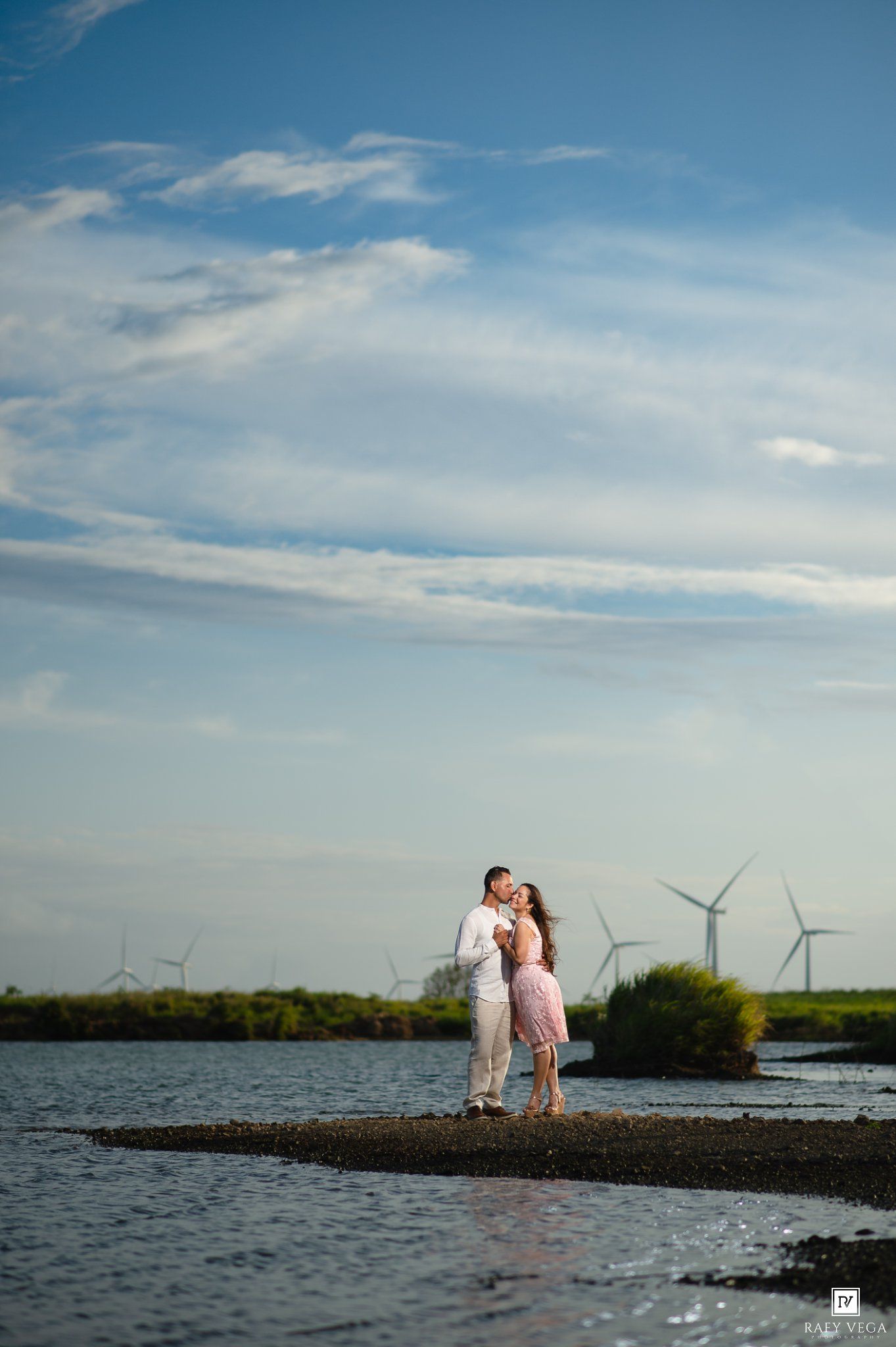 Couple embracing on a sandy shore with wind turbines and blue sky backdrop.