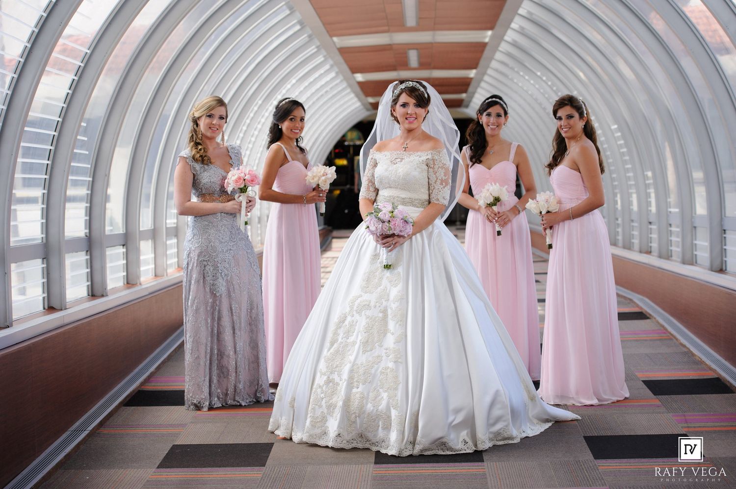Bride in white ball gown poses with four bridesmaids in pink dresses inside an elegant glass tunnel walkway.