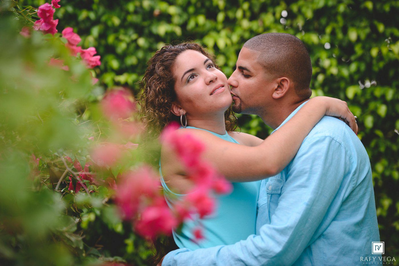 A couple shares a romantic kiss surrounded by lush green hedges and vibrant pink flowers outdoors.