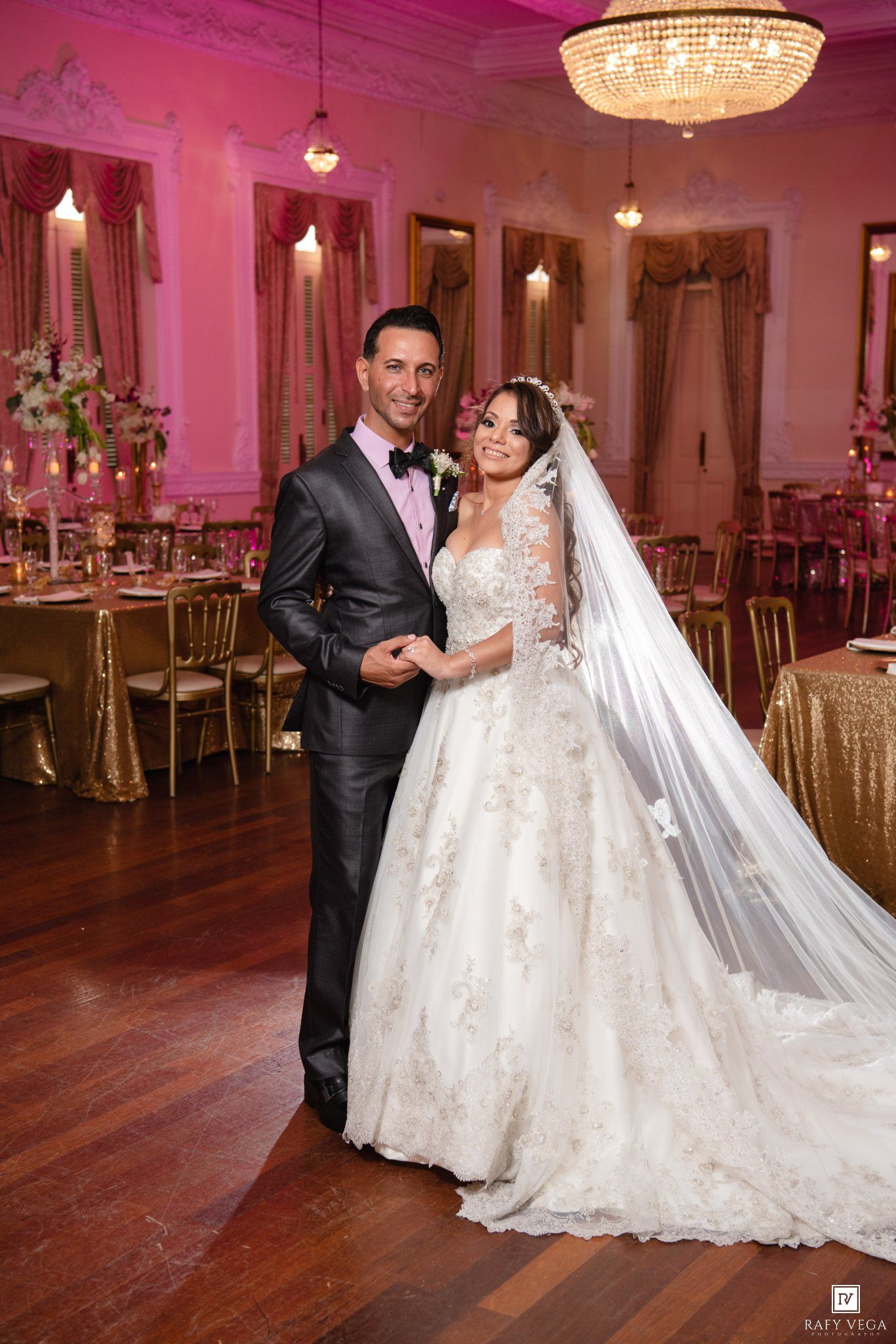 Bride in lace gown and veil poses with groom in gray tuxedo at elegant pink-lit wedding reception venue.