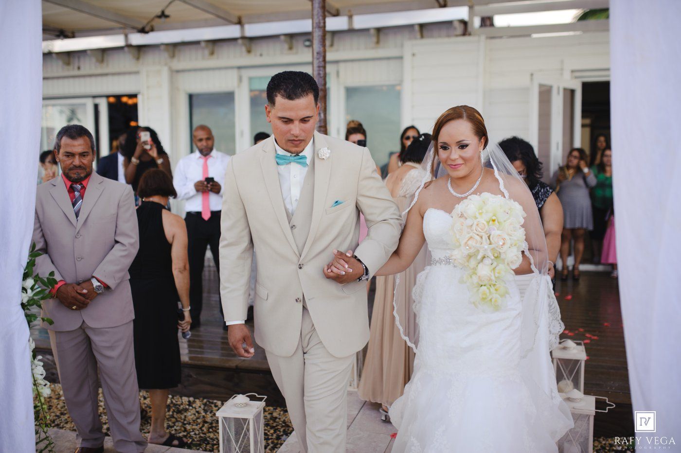 Bride and groom walk down the aisle at an outdoor wedding ceremony, groom in tan suit with teal bow tie.