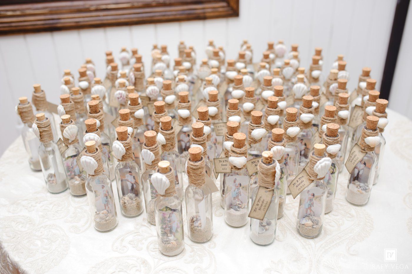 Small glass bottles with cork stoppers filled with sand and tiny photos, arranged as wedding favors on a white table.