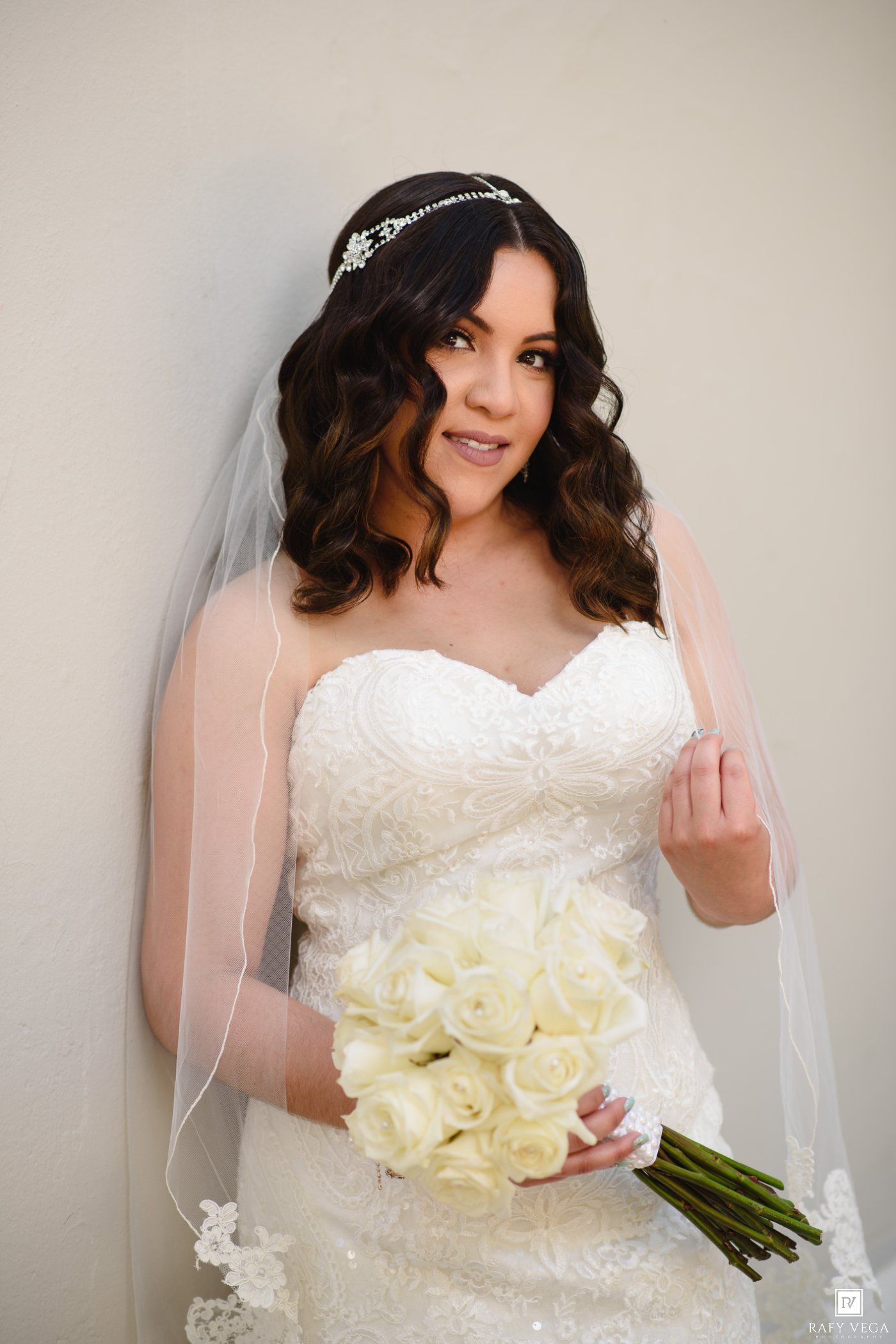 Bride in white lace strapless gown holding white rose bouquet, wearing veil and crystal headpiece, posing against neutral wall.