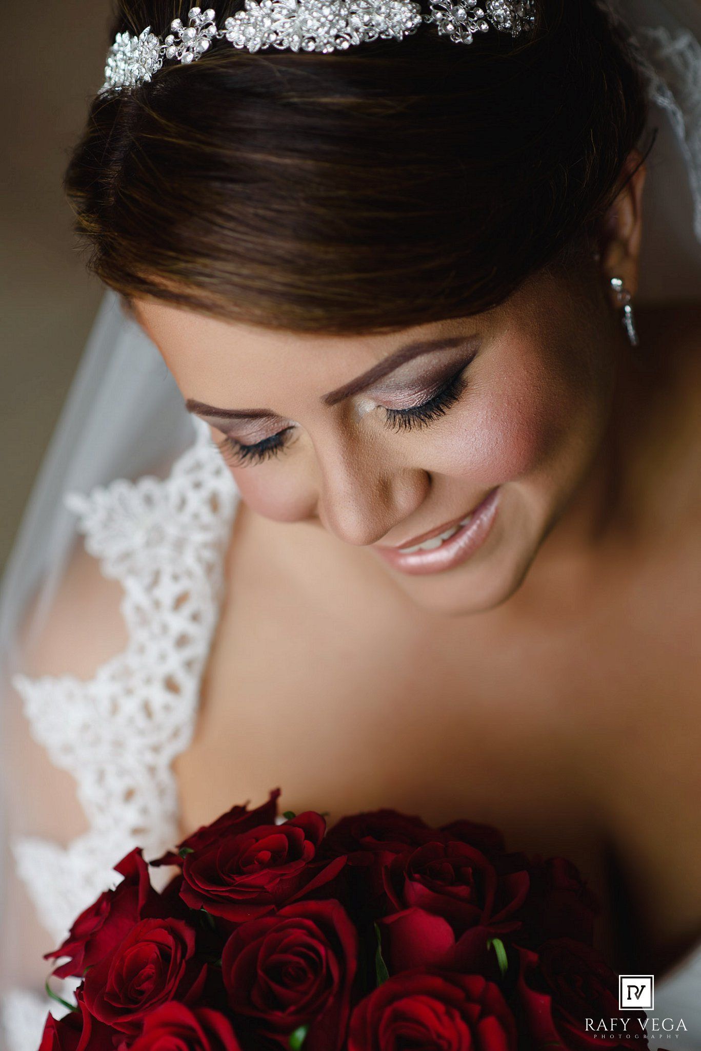 Smiling bride wearing a crystal tiara and lace gown holds a bouquet of red roses on her wedding day.