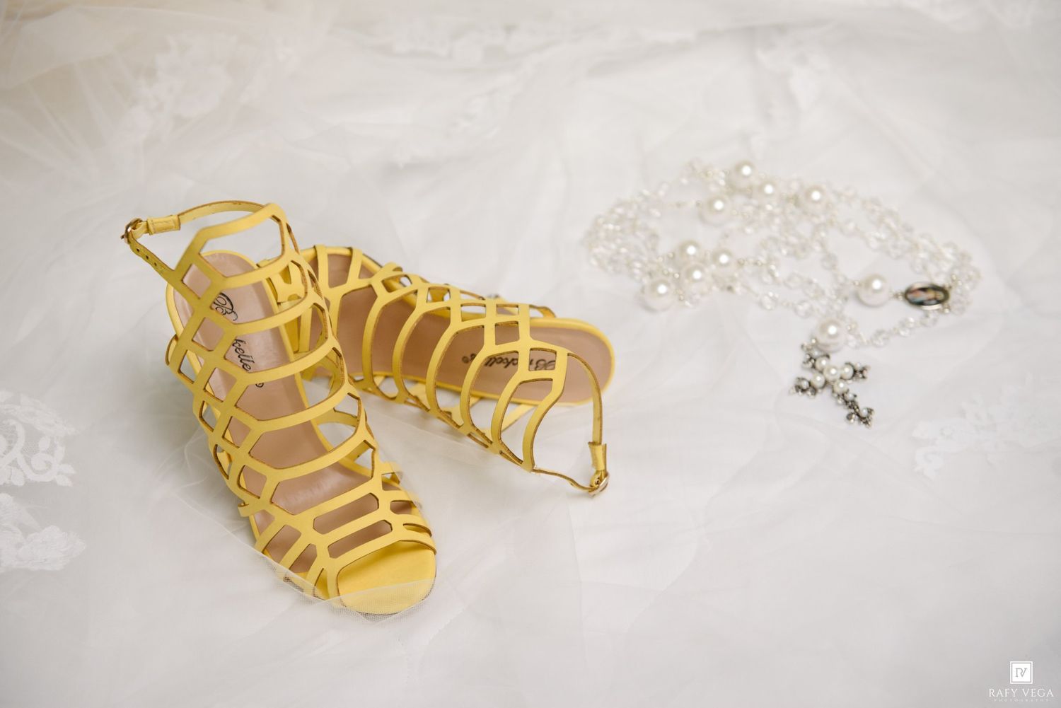 Yellow gladiator sandals beside pearl necklace and cross bracelet on white lace bridal fabric.