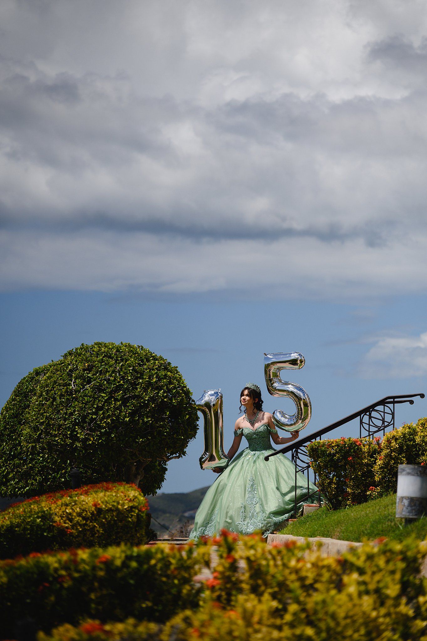 Girl in mint green quinceañera gown holds gold number 15 balloons on garden stairs under cloudy blue sky.