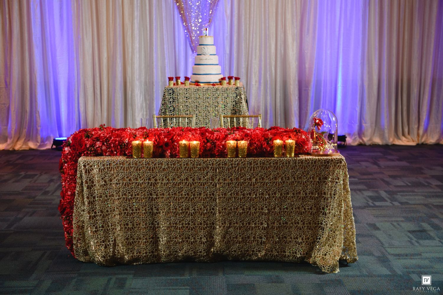Elegant wedding cake table with gold lace tablecloth, red floral arrangements, and golden candle holders in a decorated banquet hall.