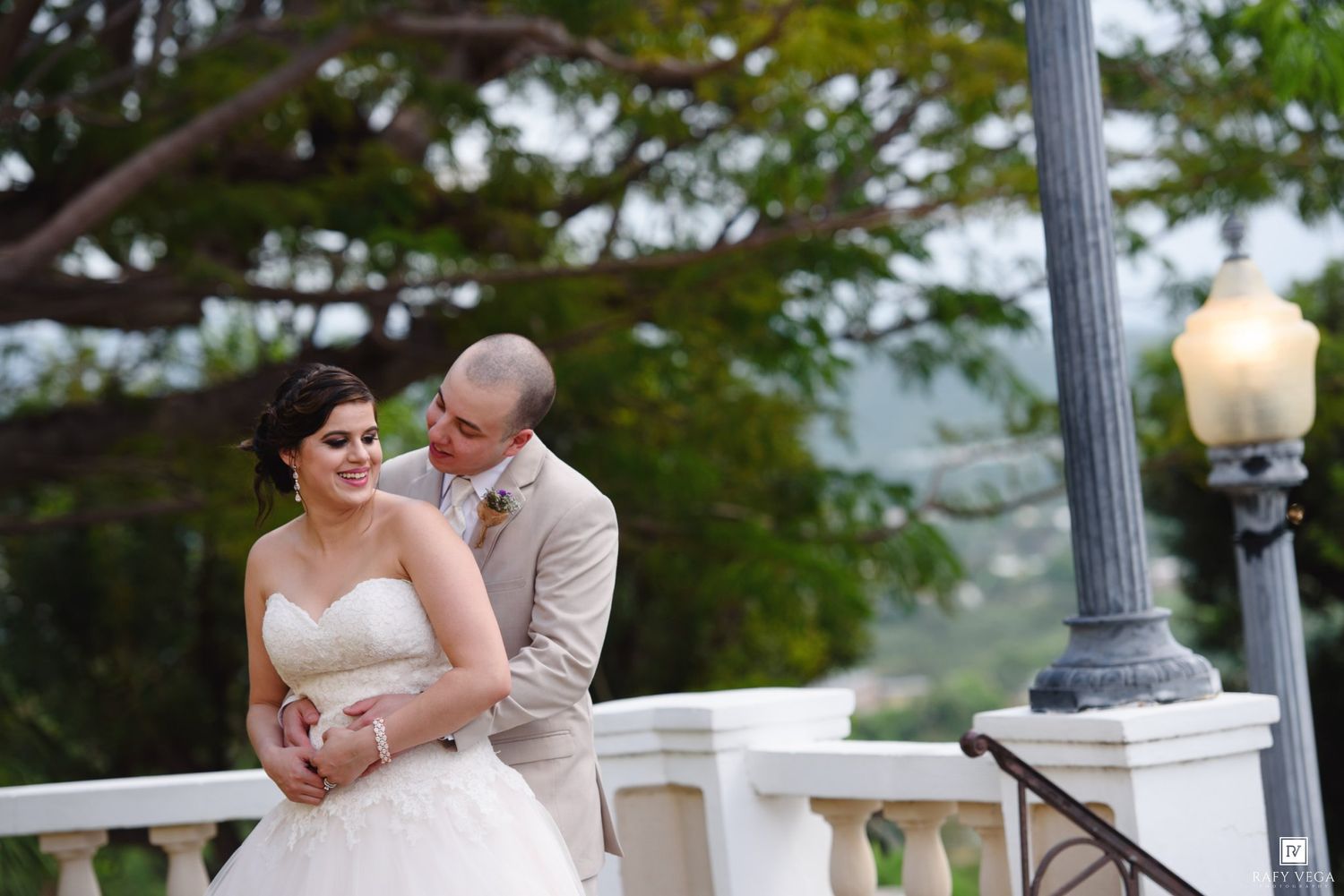 Bride and groom share a romantic moment on a white balcony, surrounded by lush greenery and a vintage street lamp.