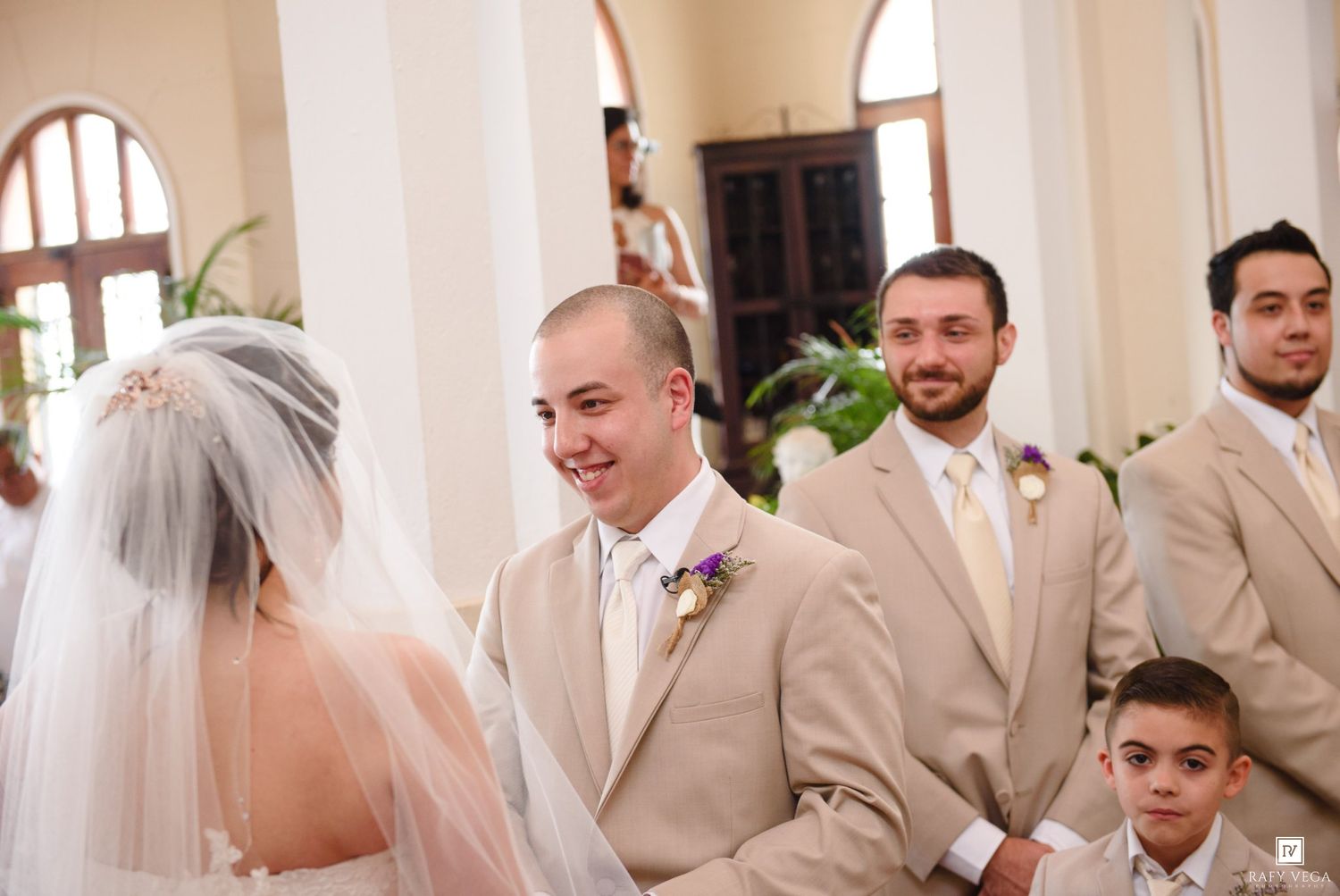 Groom smiling at bride during wedding ceremony with groomsmen in tan suits standing beside them in elegant church.