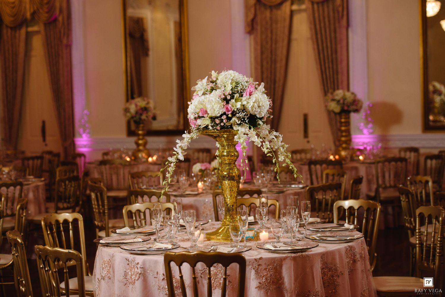 Elegant wedding reception hall with tall floral centerpieces, gold chairs, and pink candlelit tables.
