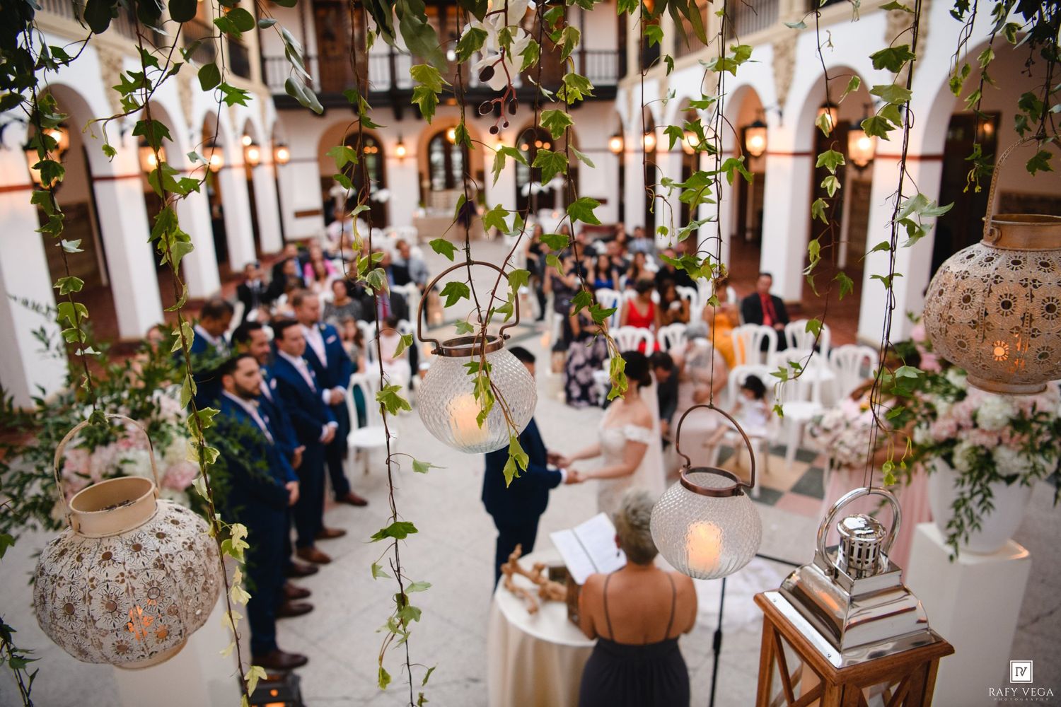 Elegant indoor wedding ceremony in a courtyard adorned with hanging greenery, lanterns, and guests seated in white chairs.