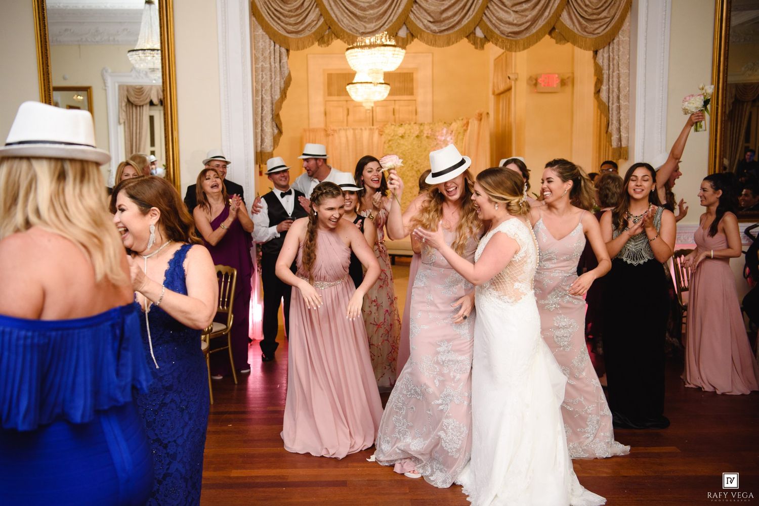 Bride and bridesmaids in pink gowns dancing joyfully at elegant wedding reception with gold chandeliers.