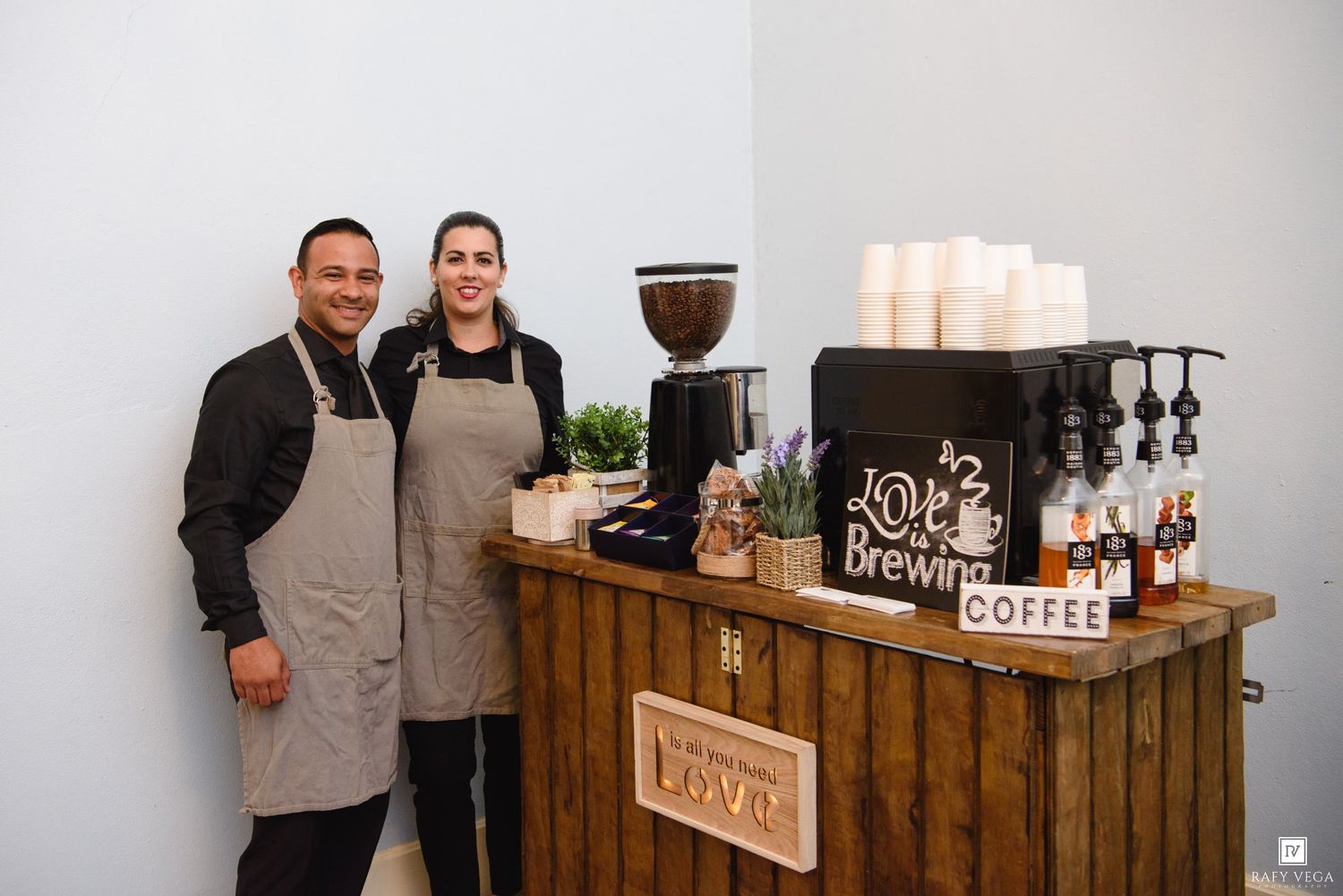 Two baristas in aprons smile behind a rustic wooden coffee bar with espresso machine and cups.