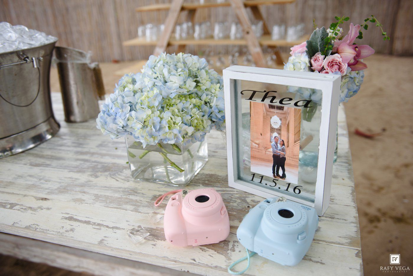 Wedding guest book table with pink and blue Instax cameras, hydrangea centerpiece, and framed couple photo dated 11.5.16.