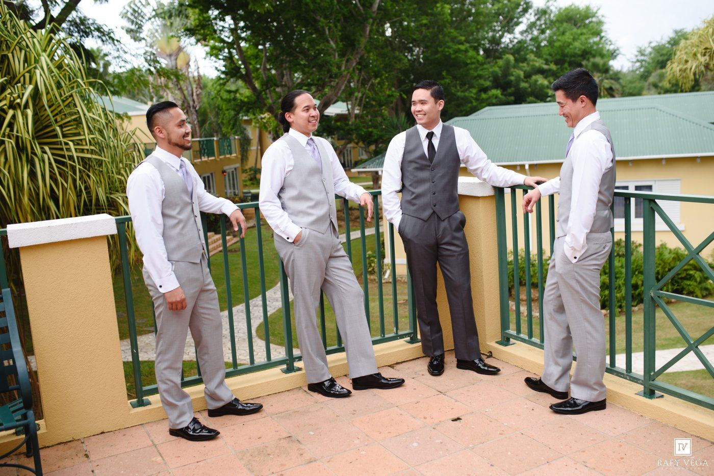 Four groomsmen in gray suits stand on a balcony outdoors, conversing before a wedding ceremony.