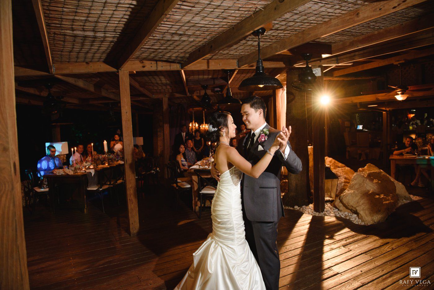 Bride and groom share their first dance in a rustic wooden venue with warm lighting and guests seated in the background.