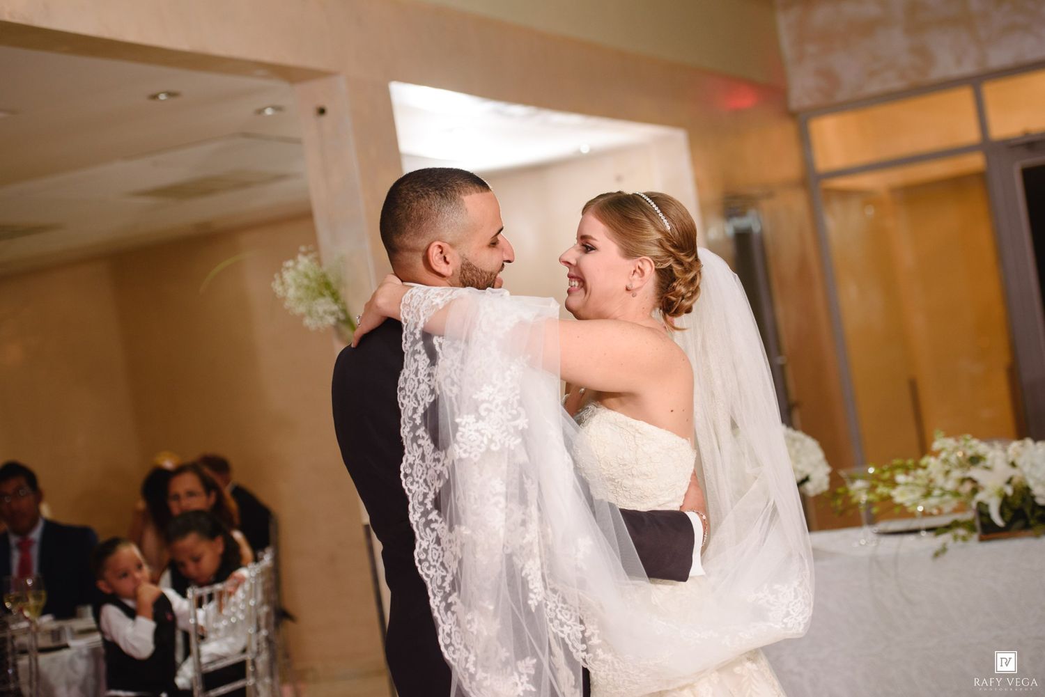 Bride and groom share their first dance, smiling joyfully, with the bride's lace veil flowing gracefully at their wedding reception.