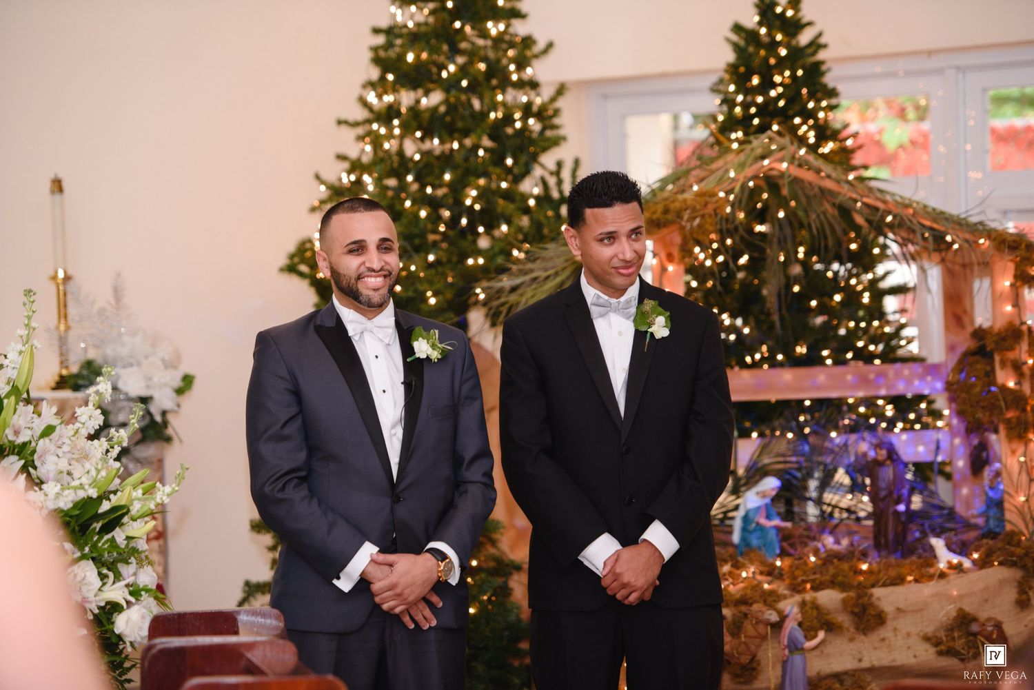 Two grooms in suits stand smiling before a decorated Christmas tree and nativity scene at a festive wedding ceremony.