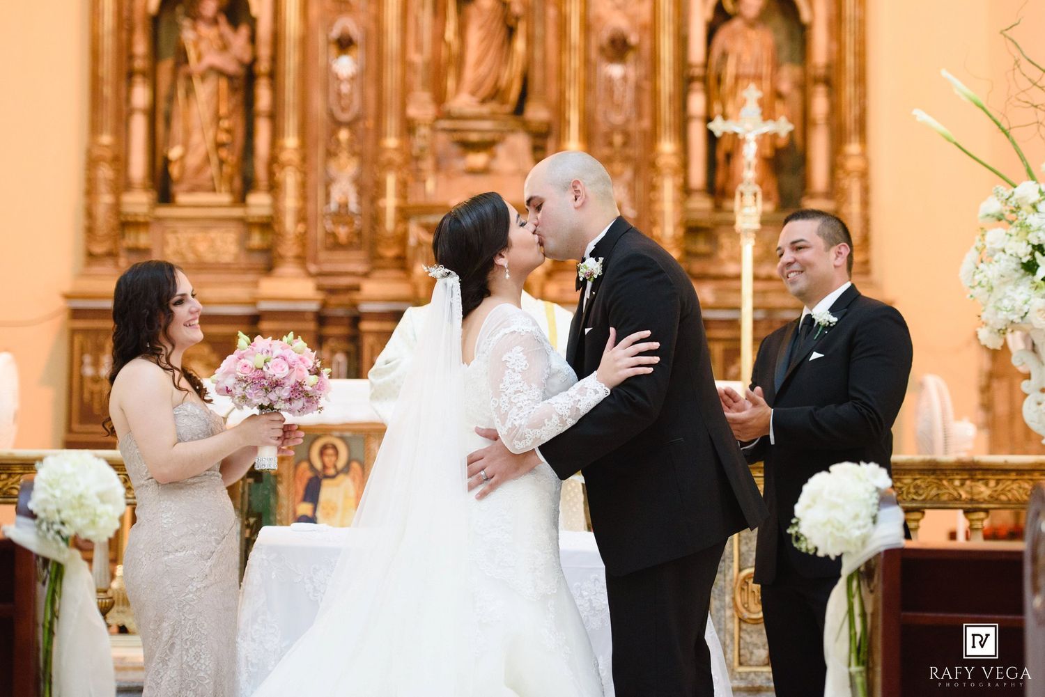 Bride and groom share first kiss at church altar during wedding ceremony with bridesmaid and officiant nearby.
