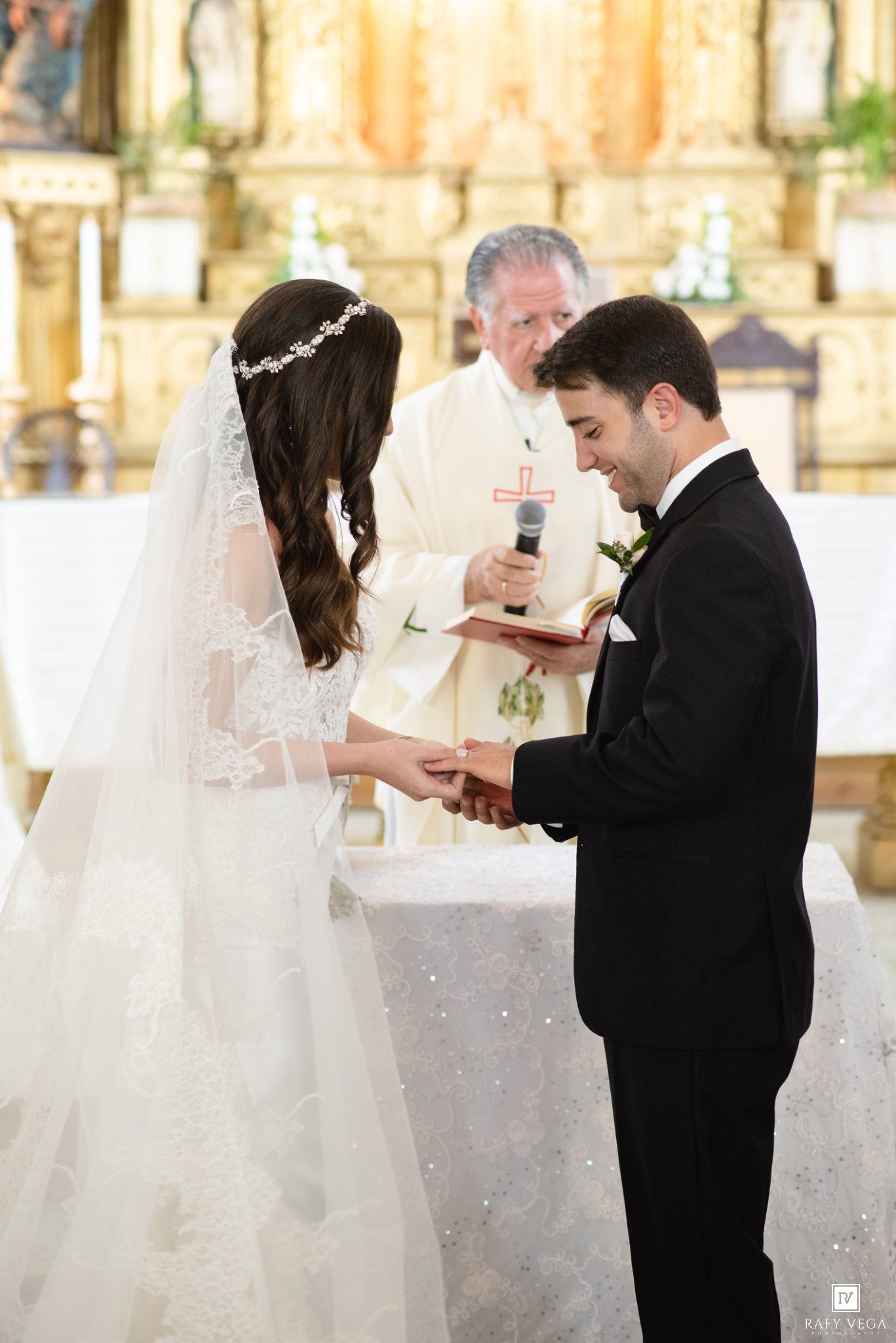 Bride and groom exchange rings at church altar during wedding ceremony with officiant holding book behind them.
