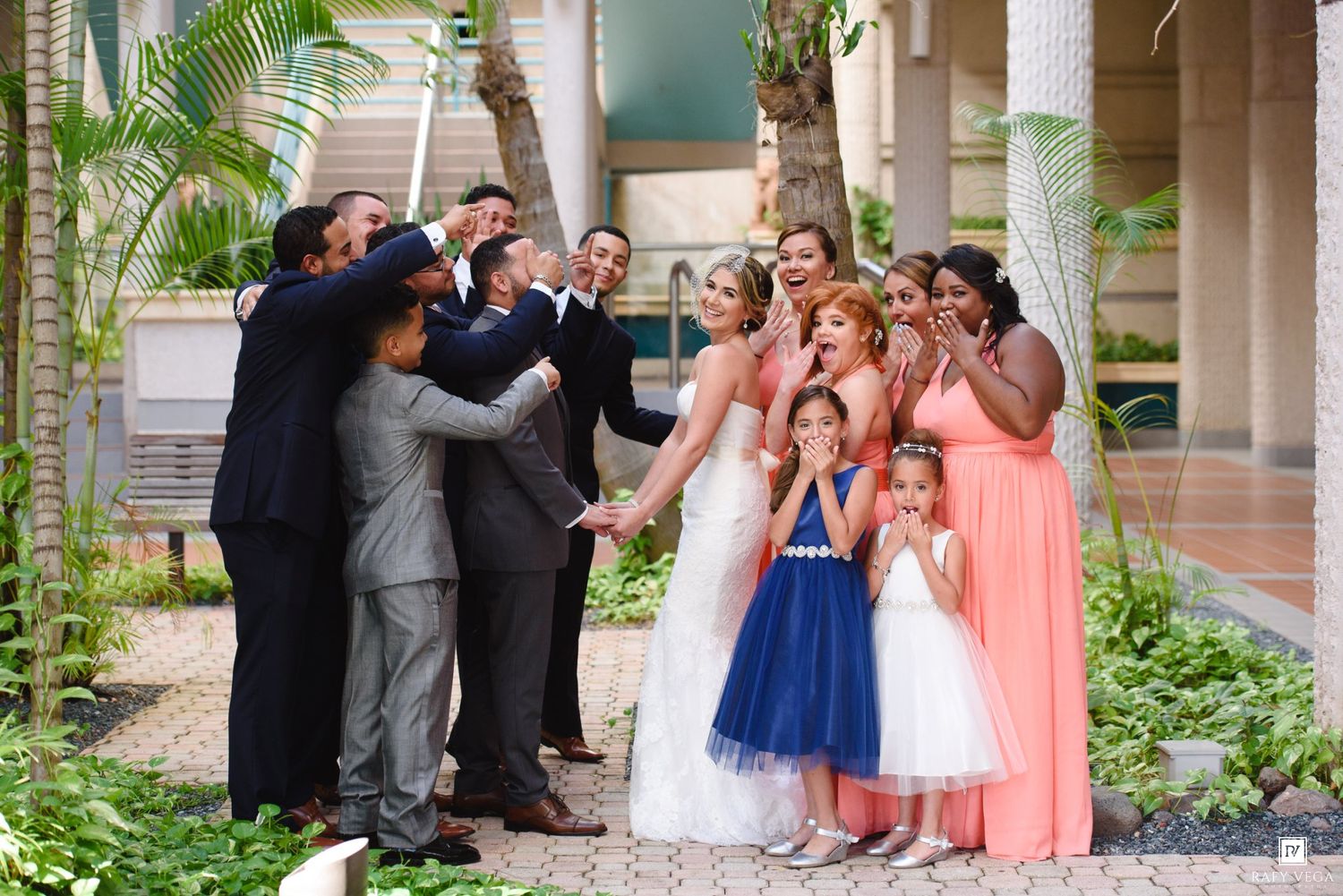 Wedding party group photo with bride, groom, bridesmaids in coral and navy, groomsmen, and flower girls in a tropical venue.