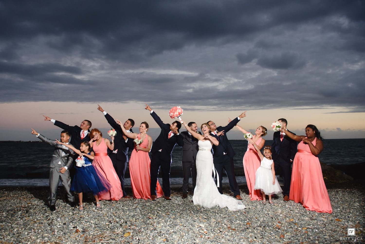 Wedding party celebrating on a rocky beach under dramatic stormy skies, bridesmaids in coral dresses cheering with arms raised.