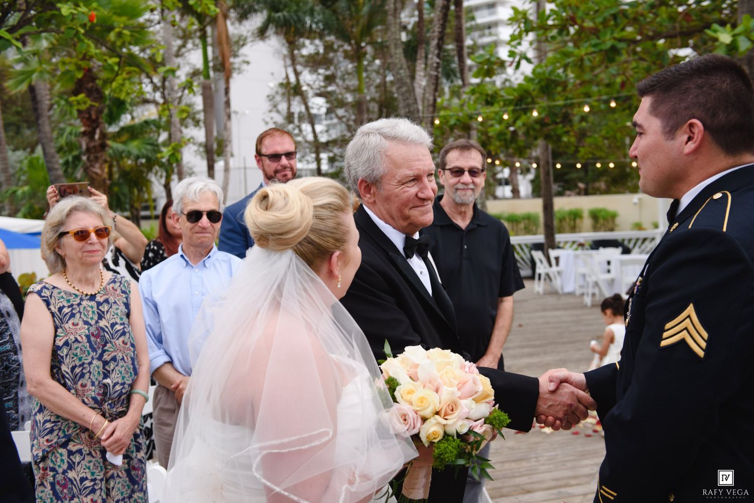 Bride in white gown and veil holding bouquet as groom in military uniform shakes hands with wedding guest outdoors.