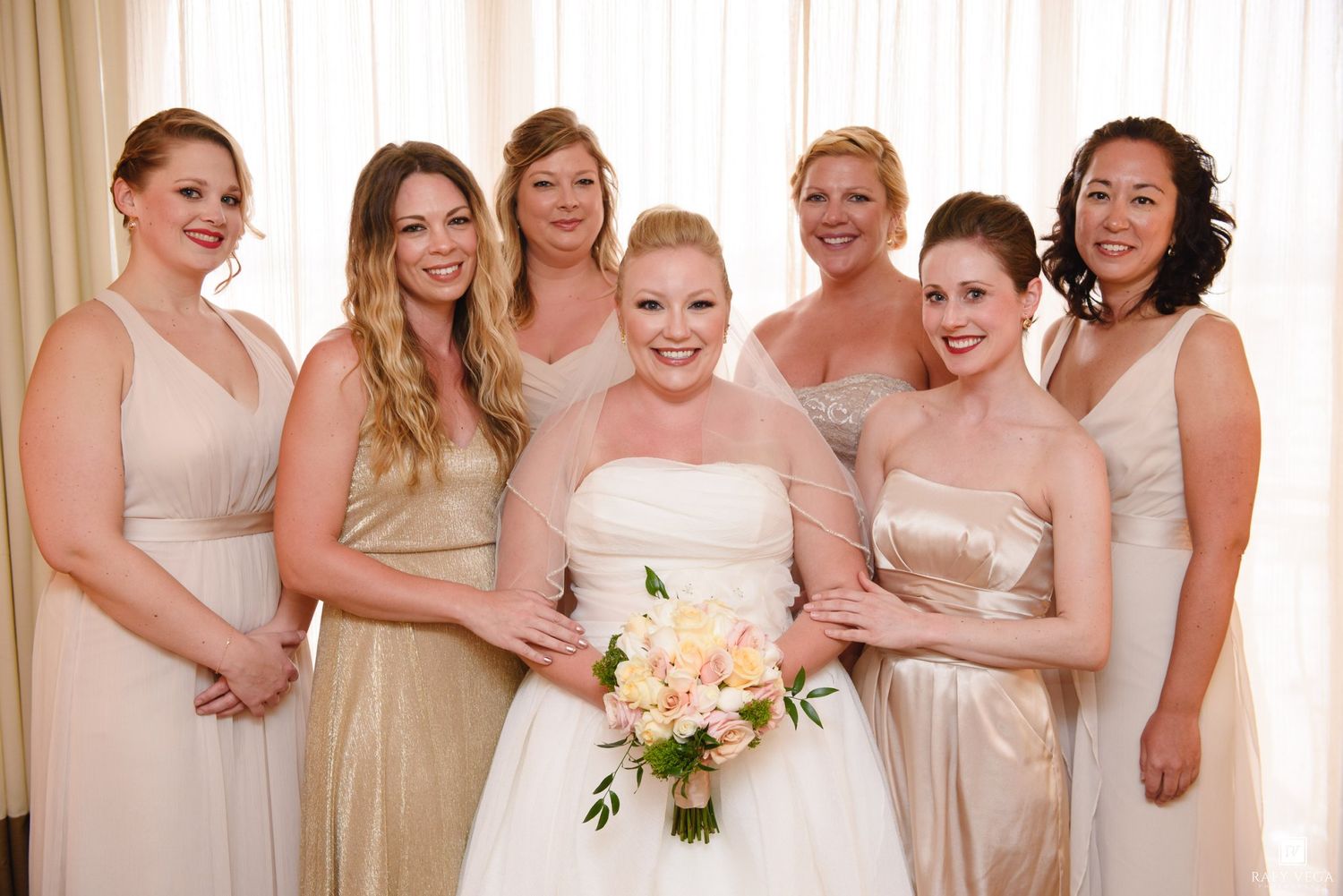 Bride in white strapless gown holds pink bouquet surrounded by bridesmaids in champagne and gold dresses.
