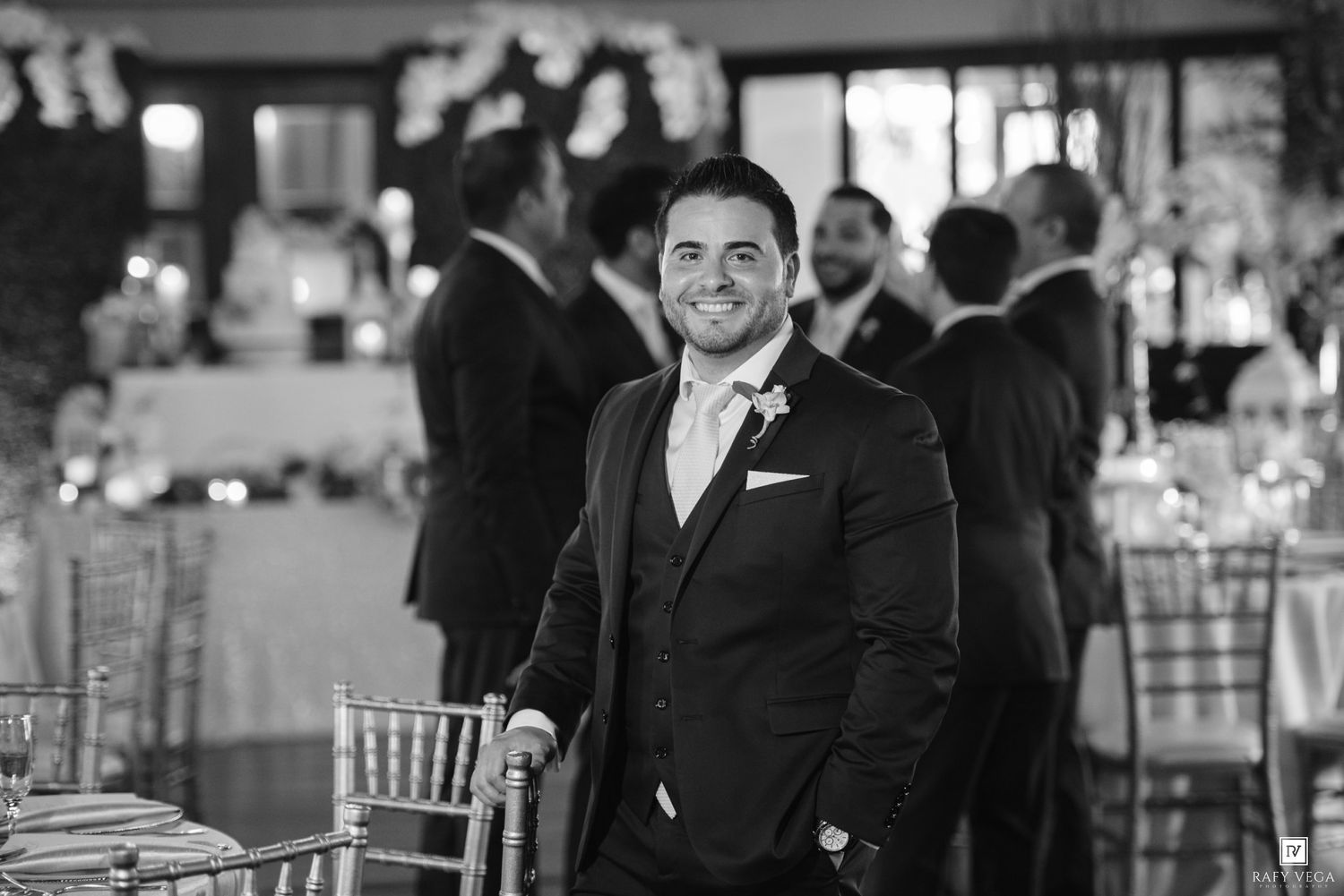 Smiling groom in dark suit with boutonniere poses at wedding reception in black and white portrait photo.