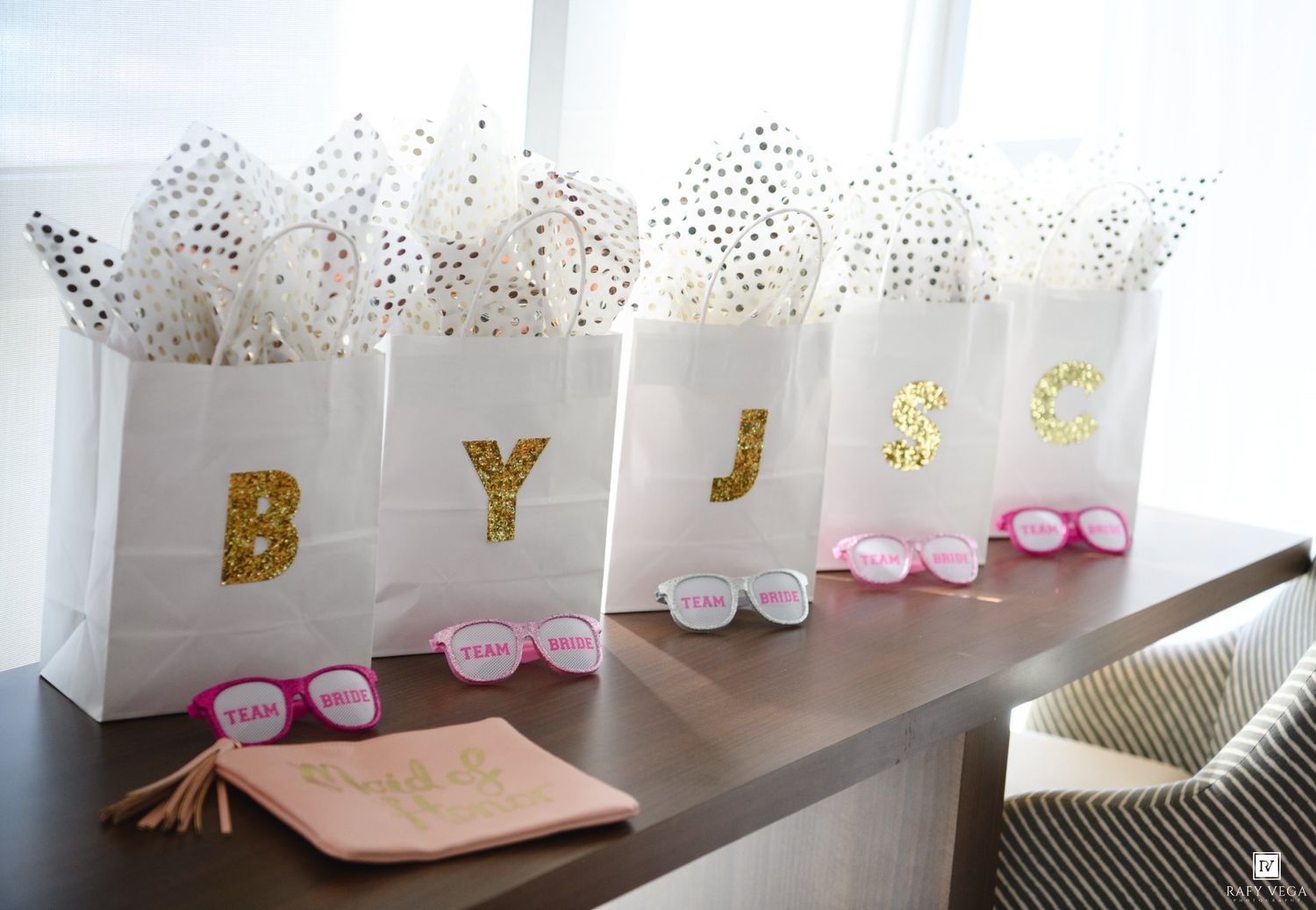 White gift bags with gold glitter monogram letters B, Y, J, S filled with tissue paper and pink sunglasses on a table.