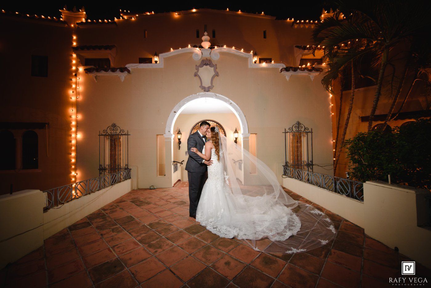 Bride and groom share romantic kiss under glowing arch with string lights at elegant outdoor wedding venue at night.