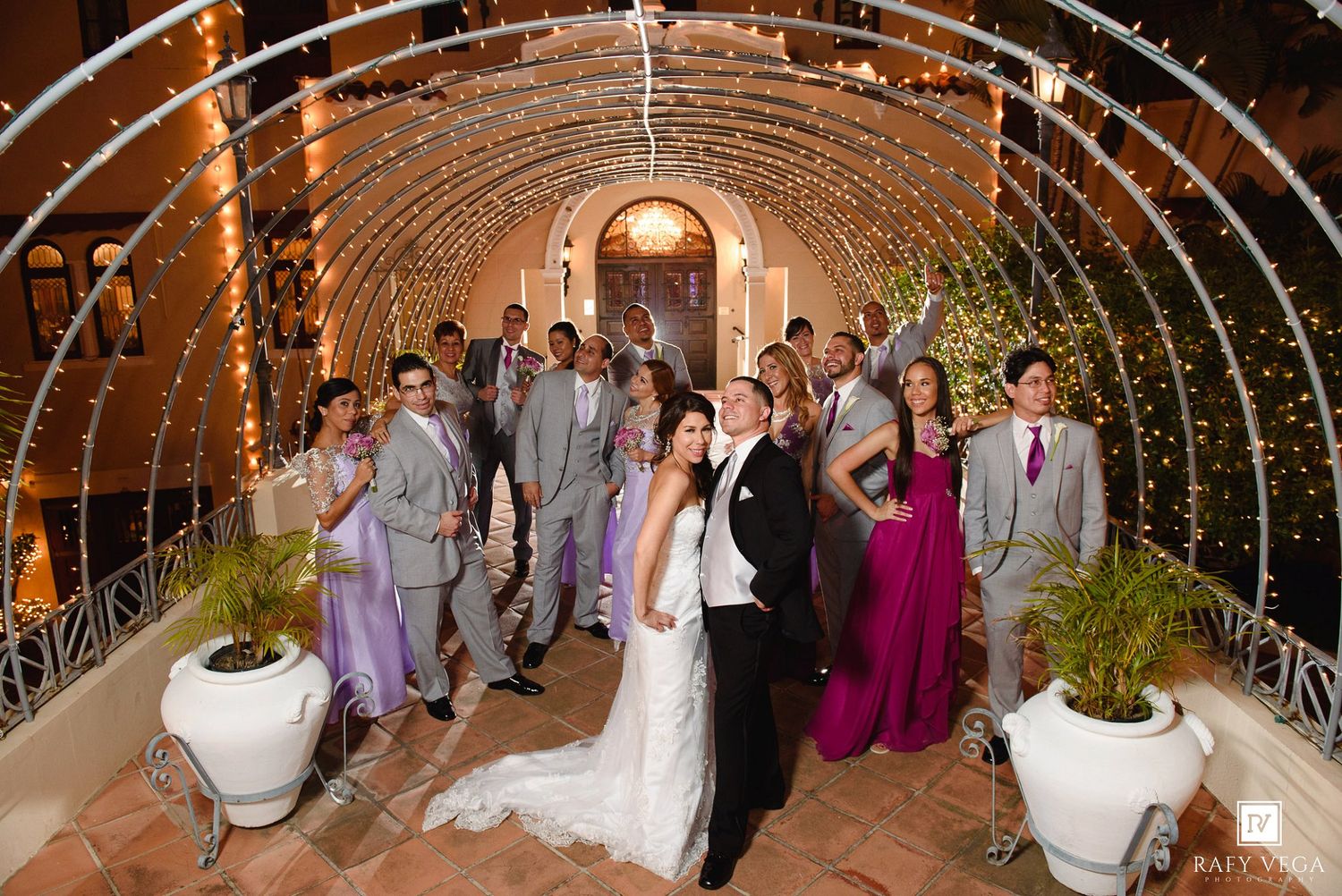 Wedding party poses under a sparkling fairy light archway at an elegant venue, bride and groom centered among guests.