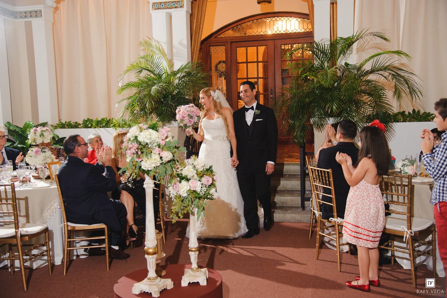 Bride and groom stand at floral altar during elegant indoor wedding ceremony with guests seated at gold chiavari chairs.