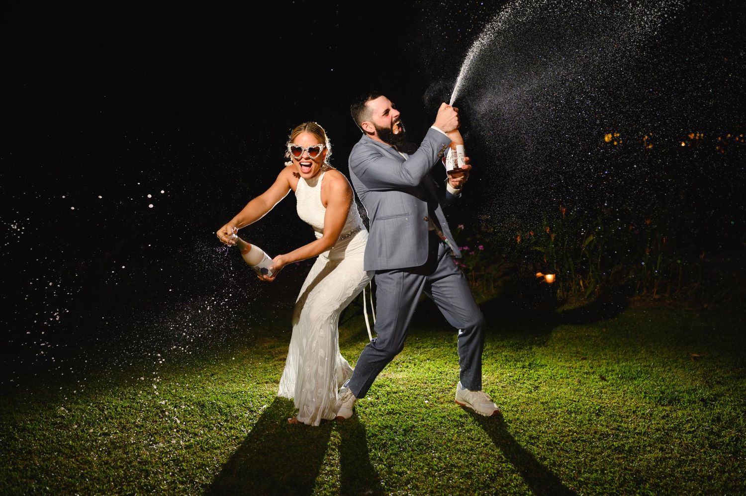 Bride and groom spraying champagne bottles at night during outdoor wedding celebration.