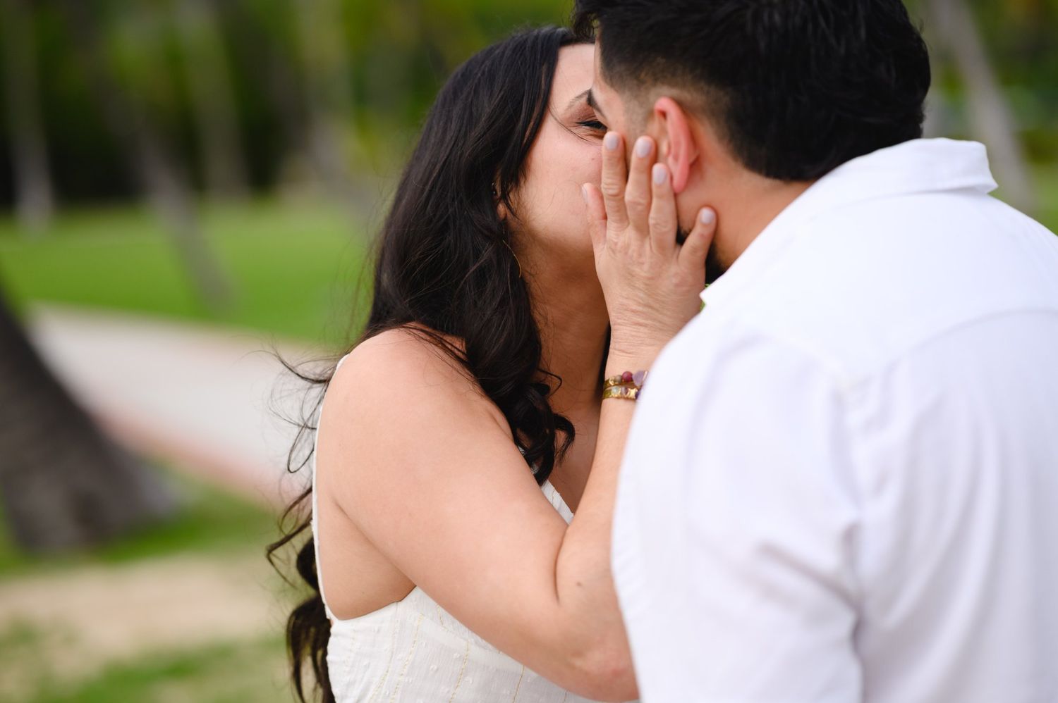 A couple shares a romantic kiss outdoors near palm trees, with the woman in a white dress touching the man's face.